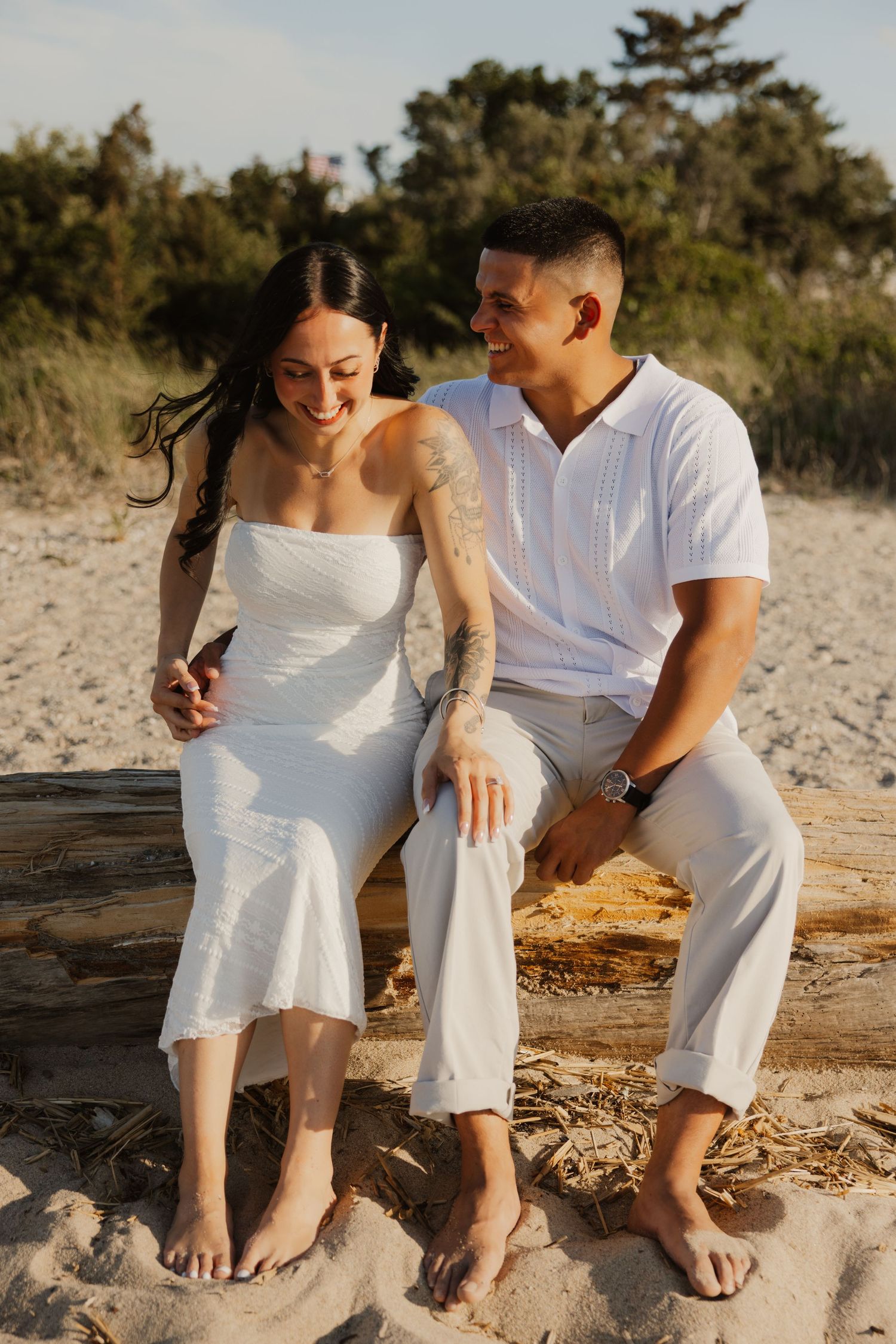 Couple sitting together on driftwood on sandy beach smiling at sunset with trees behind.