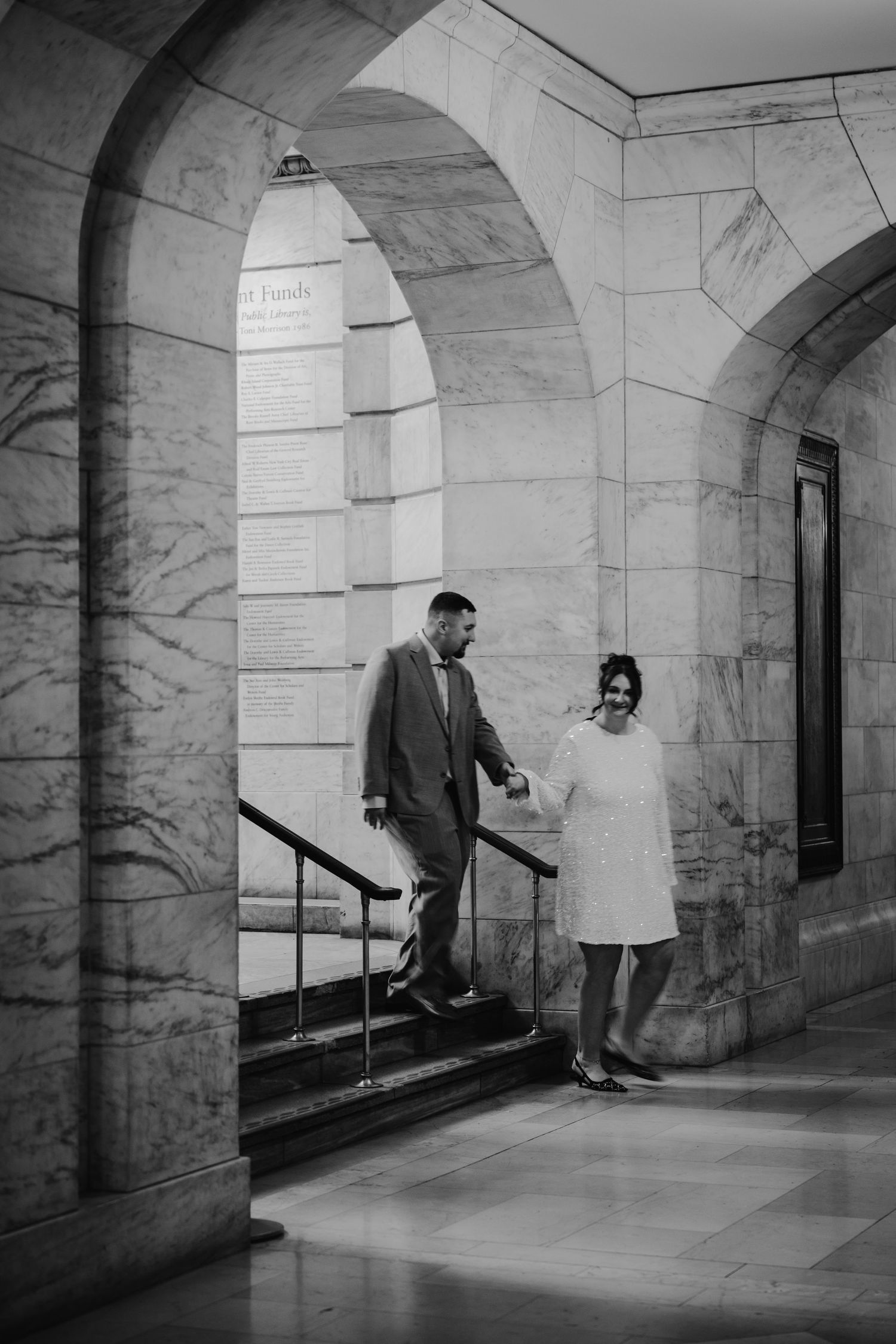 Black and white photo of couple descending marble stairs through elegant arched hallway.
