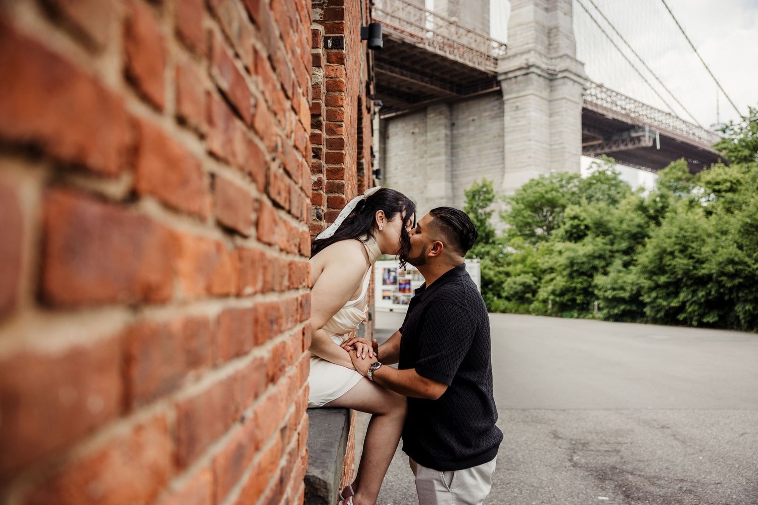 Couple sharing a kiss leaning against a brick wall with a suspension bridge behind them.