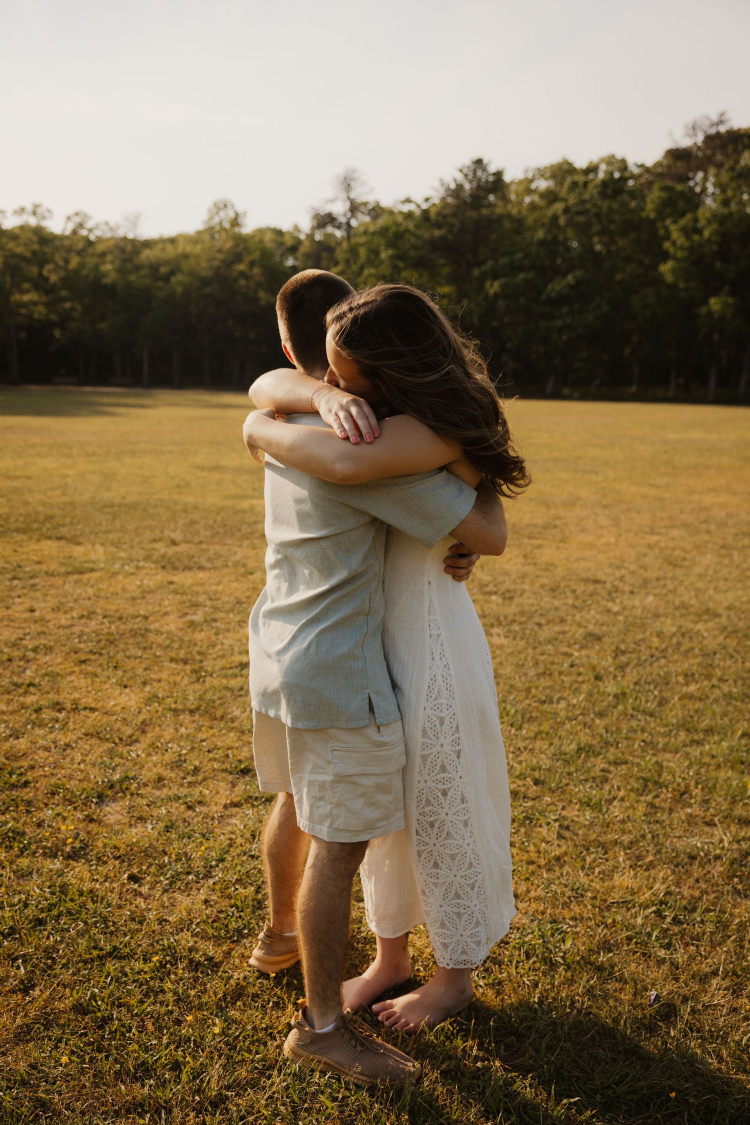 A couple shares a tender full embrace while standing barefoot in a golden sunlit grassy field.