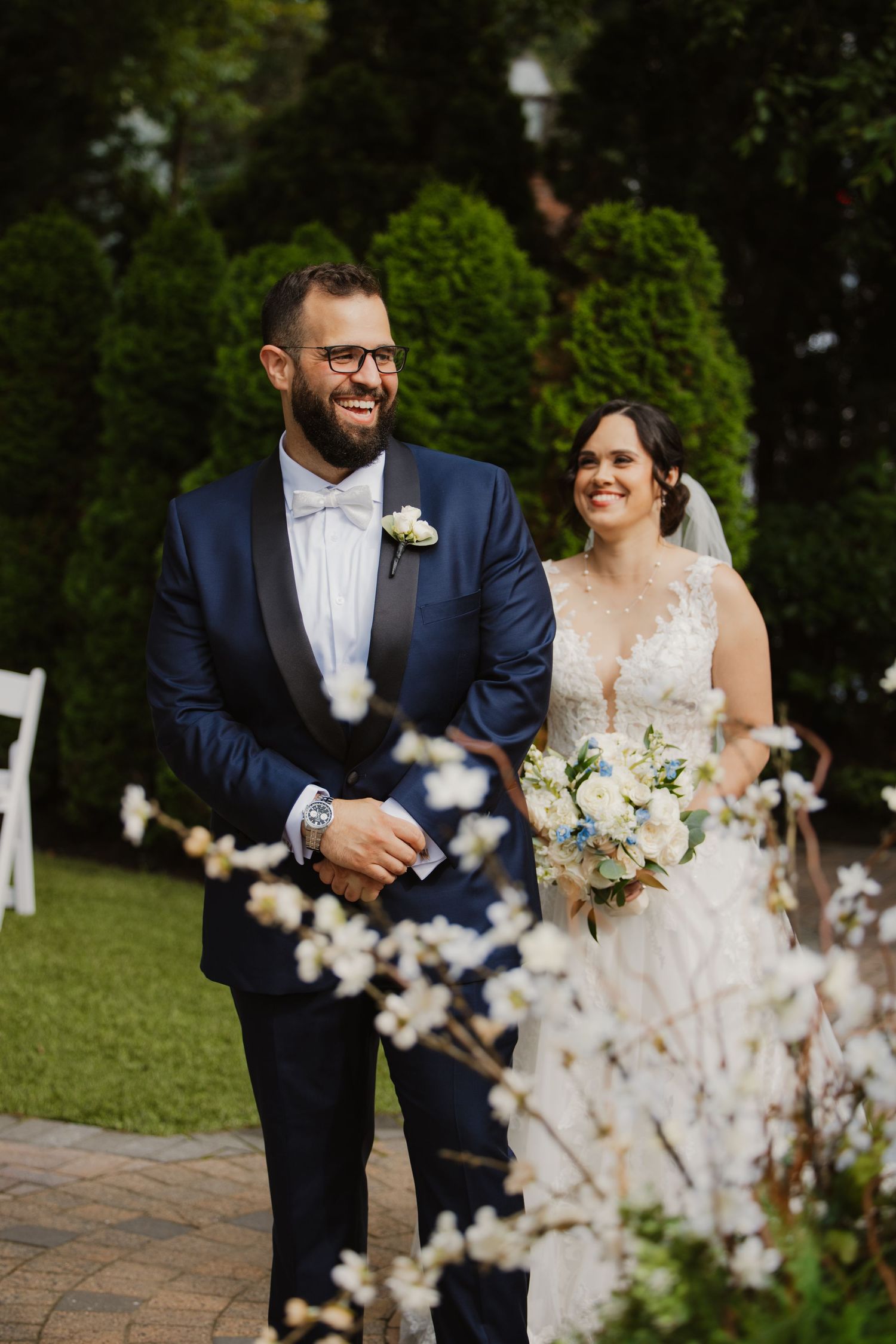 Smiling groom in navy tuxedo walks with bride holding white bouquet through garden ceremony.