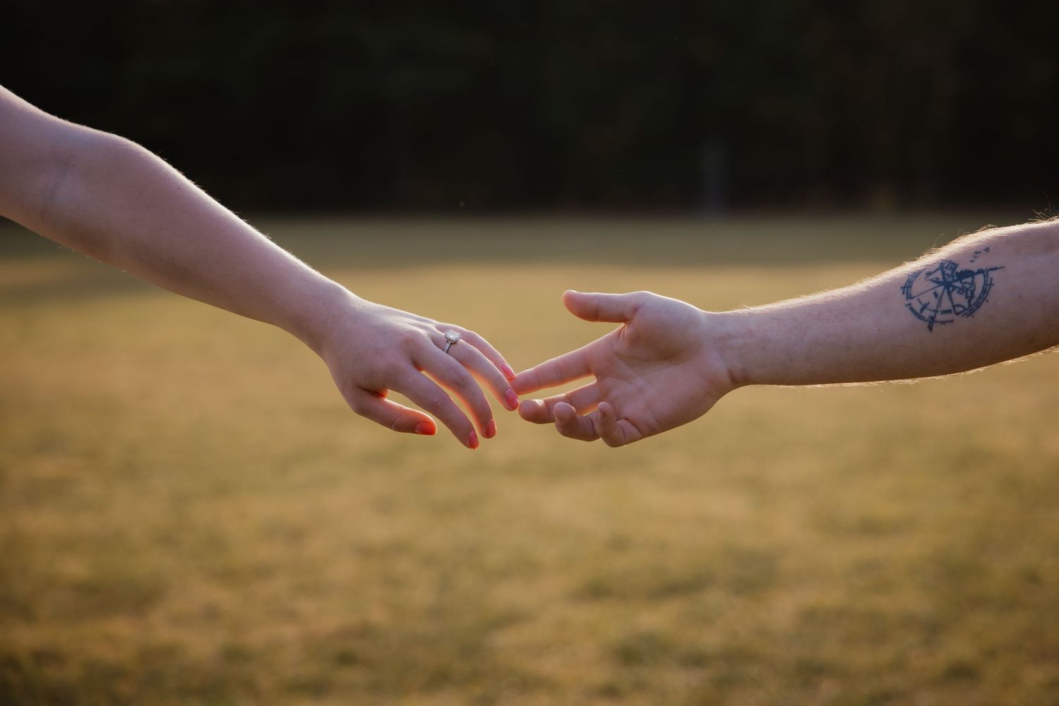 Two hands reaching toward each other with engagement ring visible and tattoo on wrist outdoors.