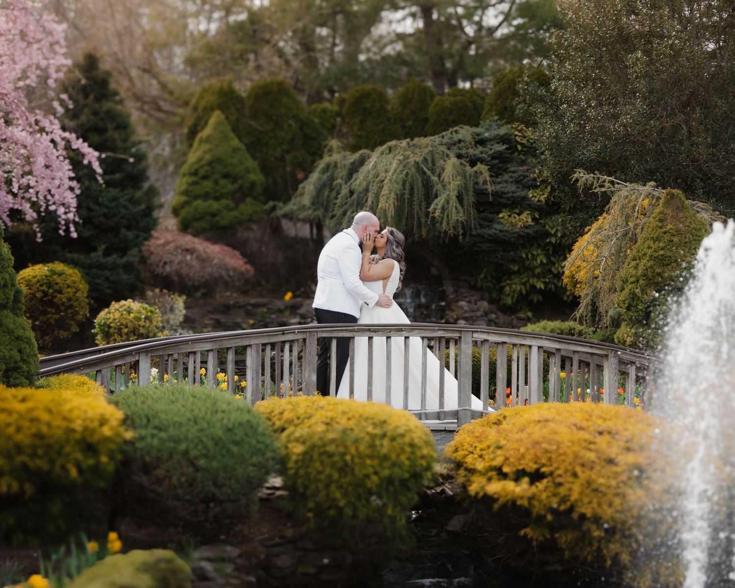 Bride and groom kissing on a white garden bridge surrounded by colorful spring flowers and a fountain.