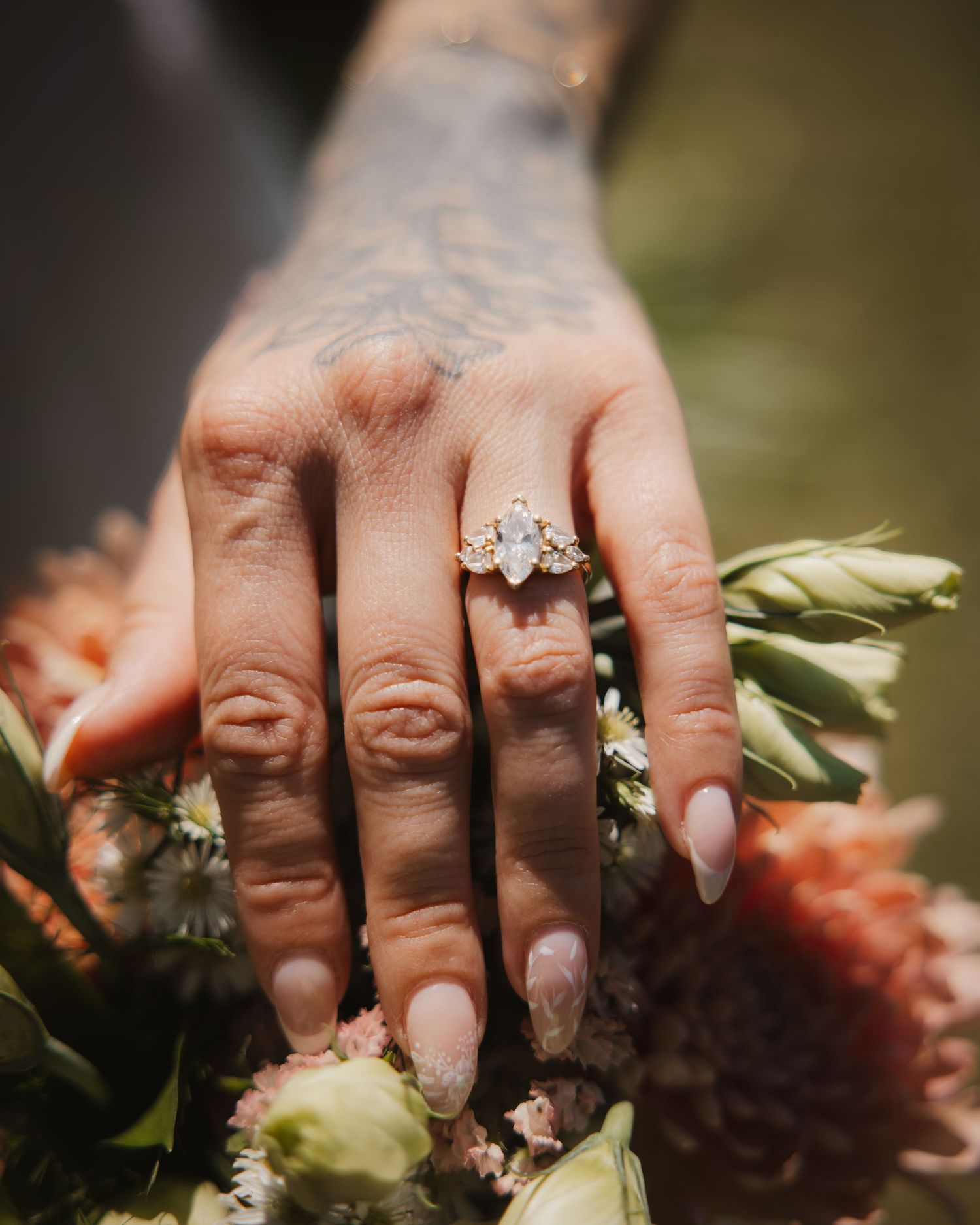 Close-up of a tattooed hand wearing a marquise gold engagement ring over a floral bouquet.