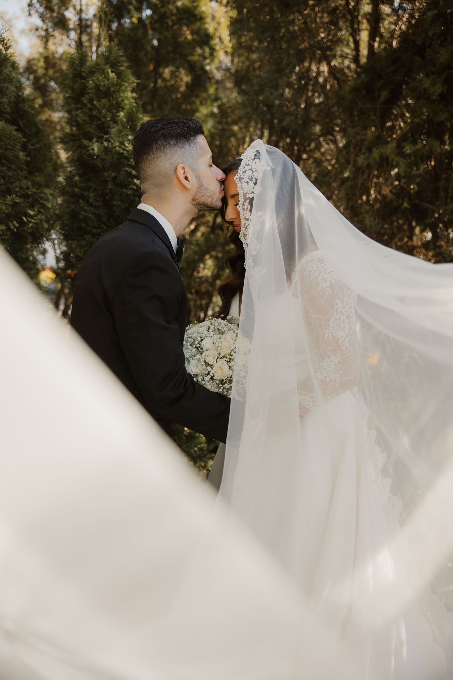 Bride and groom sharing a romantic kiss outdoors with flowing white veil and bridal bouquet.