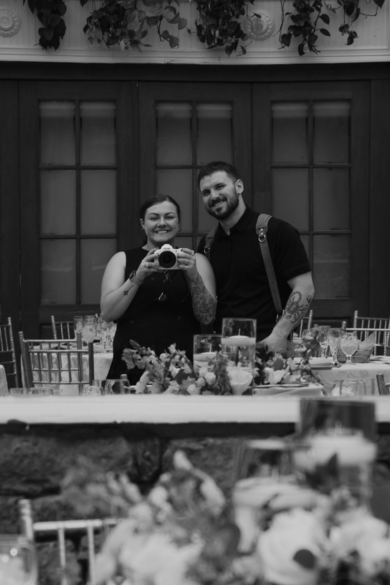 Black and white photo of two wedding photographers smiling at a decorated reception venue.