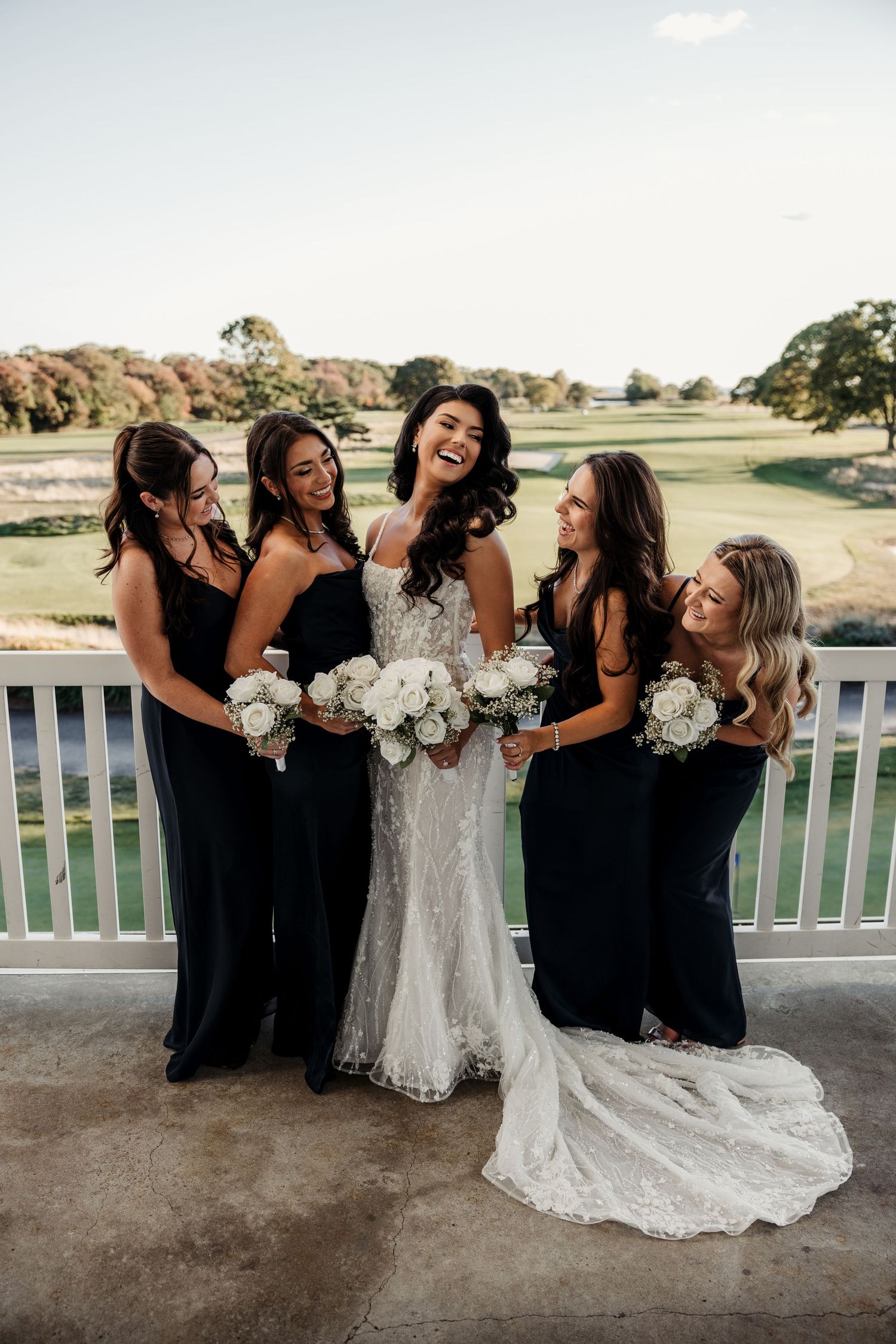 Bride in lace gown laughs with four bridesmaids in navy dresses holding white bouquets on balcony.