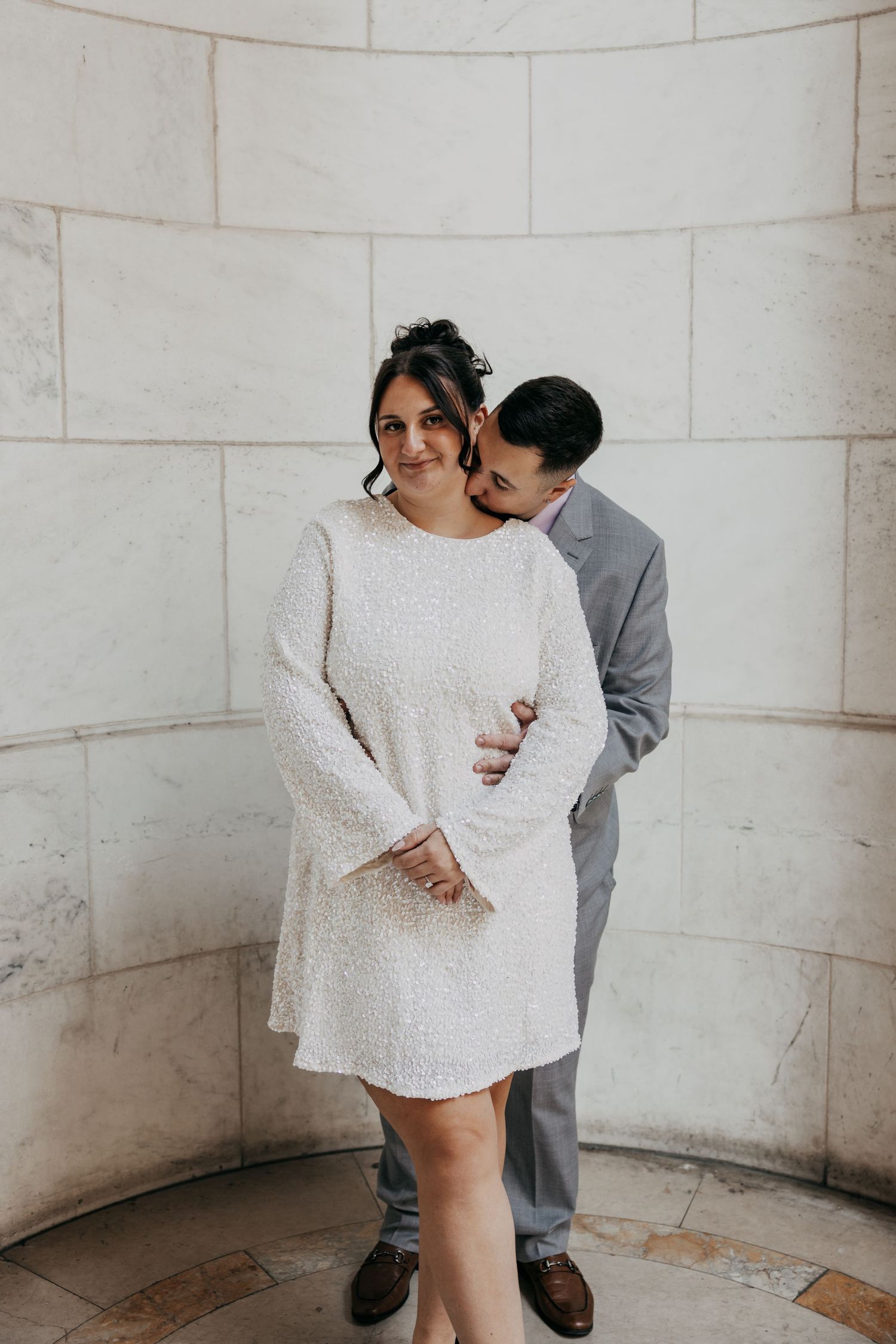 Groom embraces bride from behind in white sequin mini dress against curved marble wall.