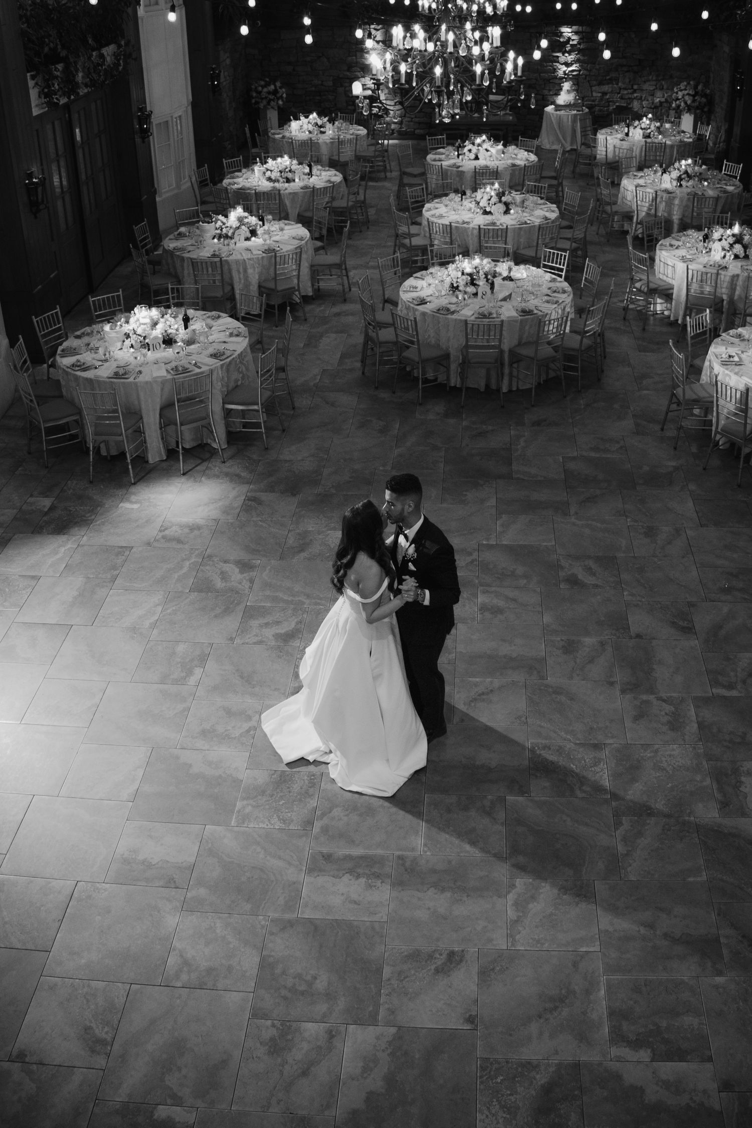 Aerial black and white photo of bride and groom sharing first dance on stone floor at elegant wedding reception.