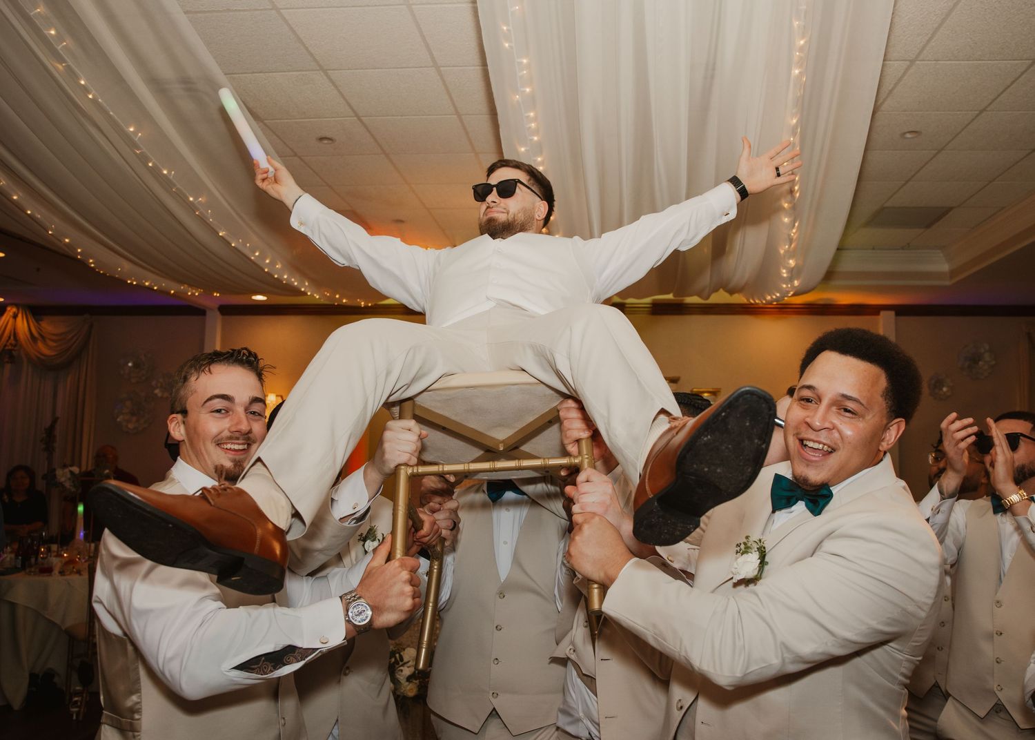 Groomsmen lifting groom on chair during lively wedding reception celebration.