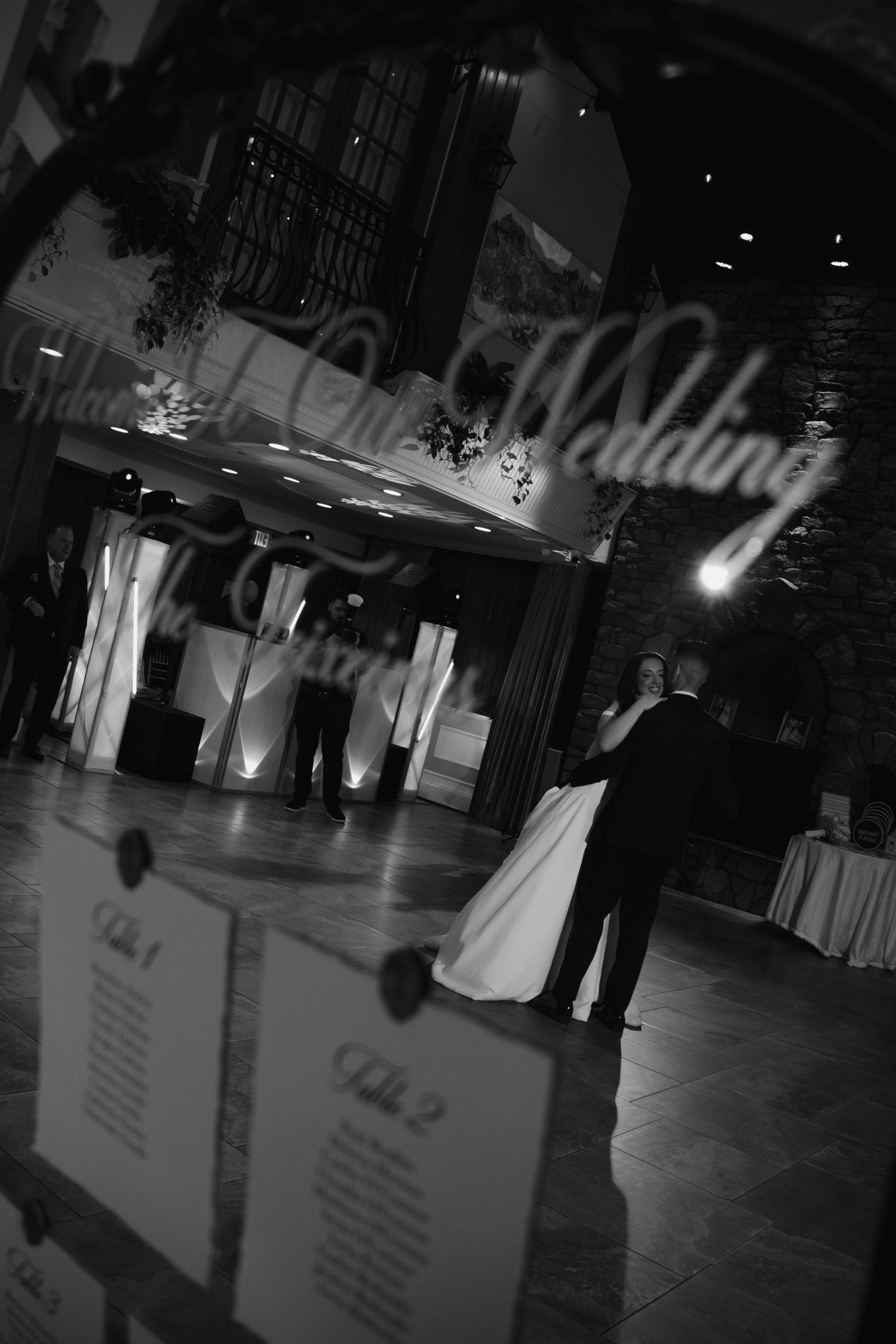 Black and white wedding reception photo with couple dancing under Our Wedding sign reflection.