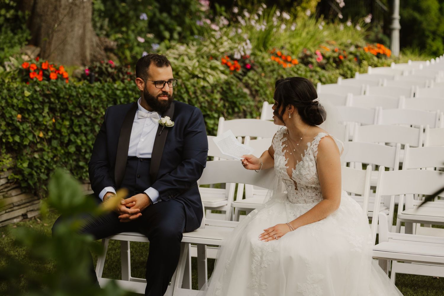 Bride and groom share an intimate moment seated on white chairs amid colorful garden flowers.