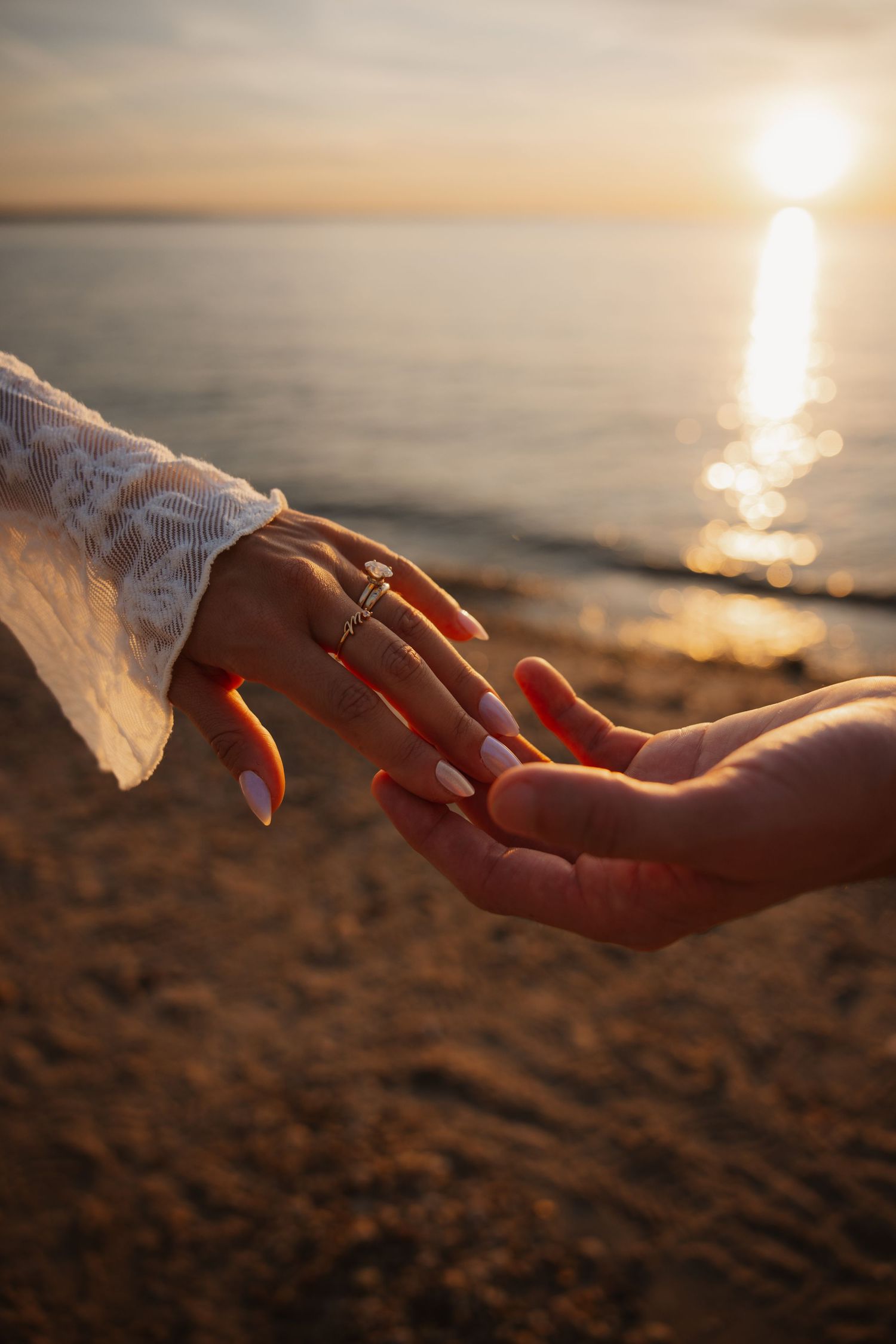 Couple holding hands on a sandy beach at sunset, woman wearing an engagement ring and white lace sleeve.