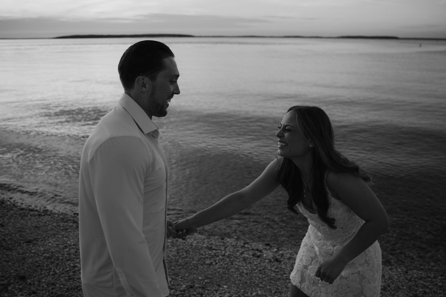 Black and white photo of a couple sharing a romantic moment on a rocky beach at sunset near calm waters.
