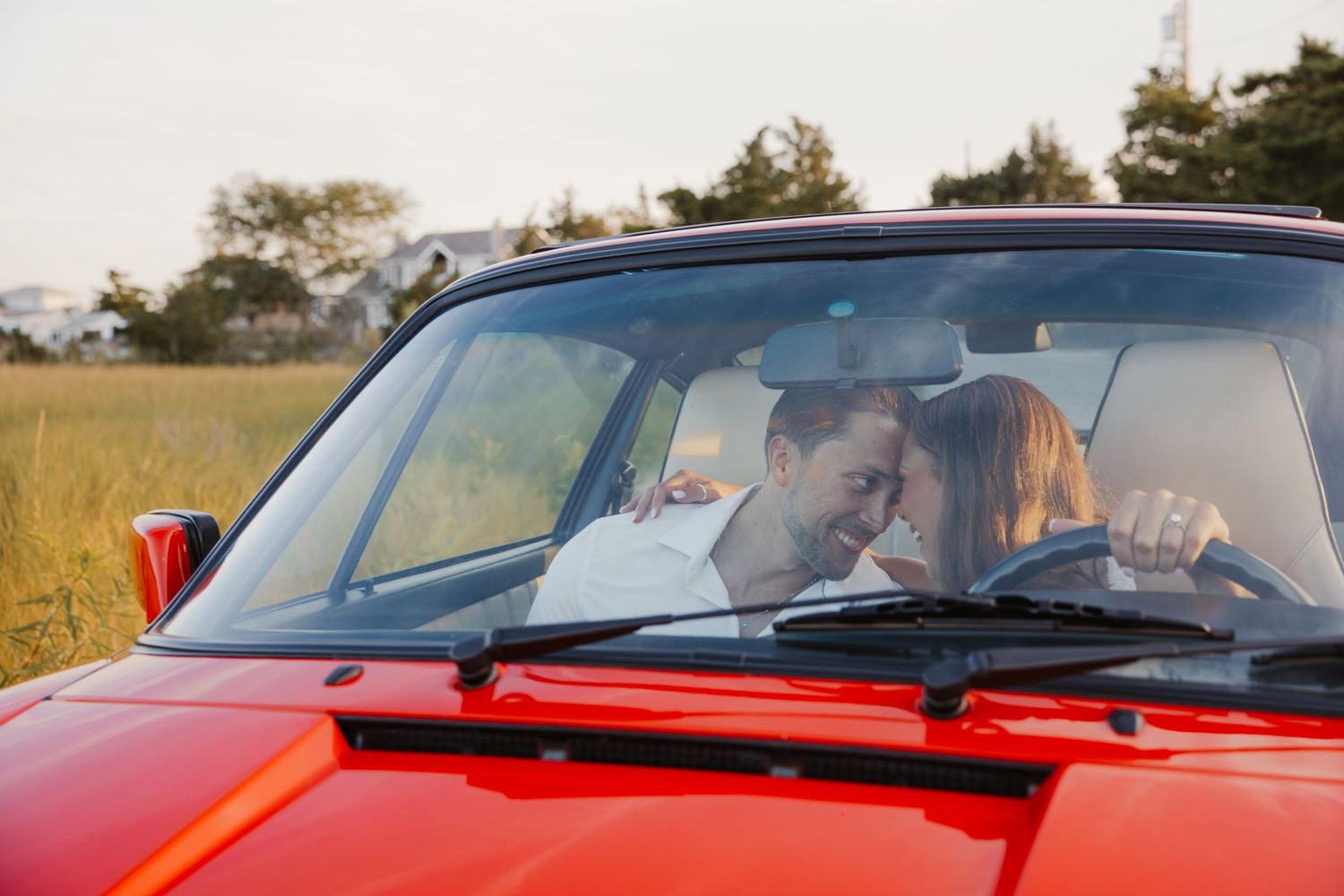 Couple sharing a romantic moment in a red convertible car parked near a green field at sunset.