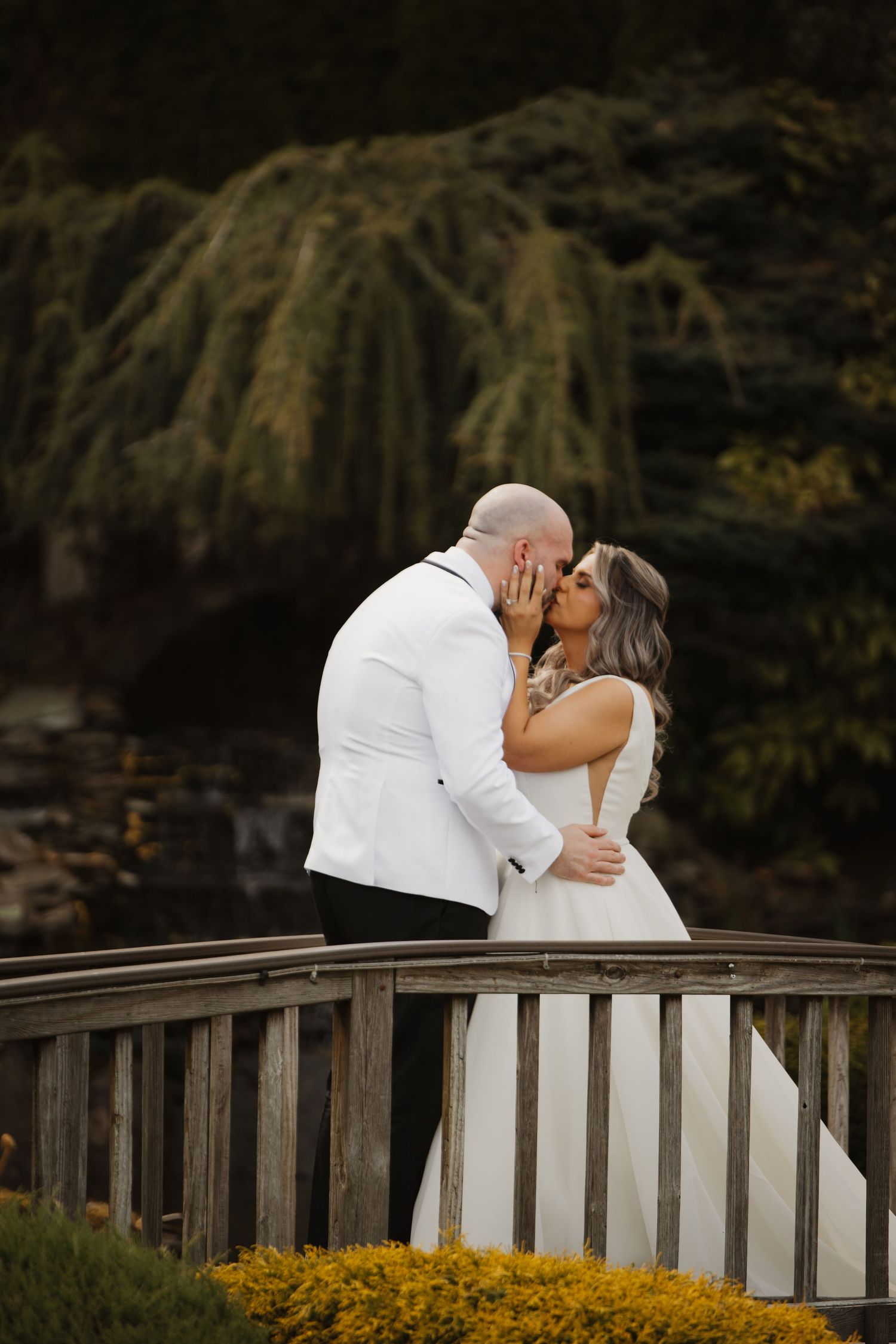 A bride and groom share a romantic kiss on a wooden bridge surrounded by lush greenery on their wedding day.