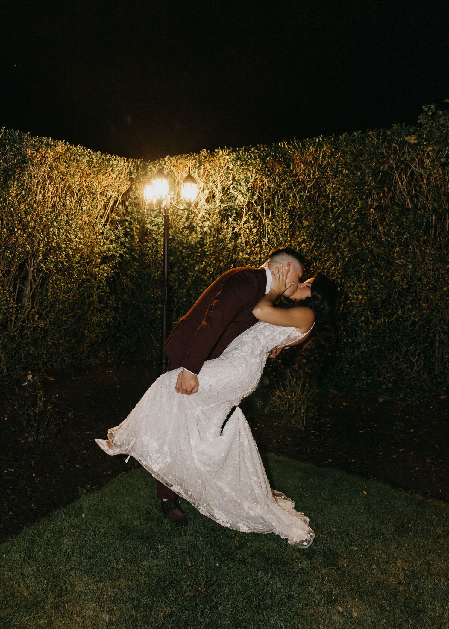 Groom in burgundy suit dips bride in lace gown for a kiss under garden lights at night.