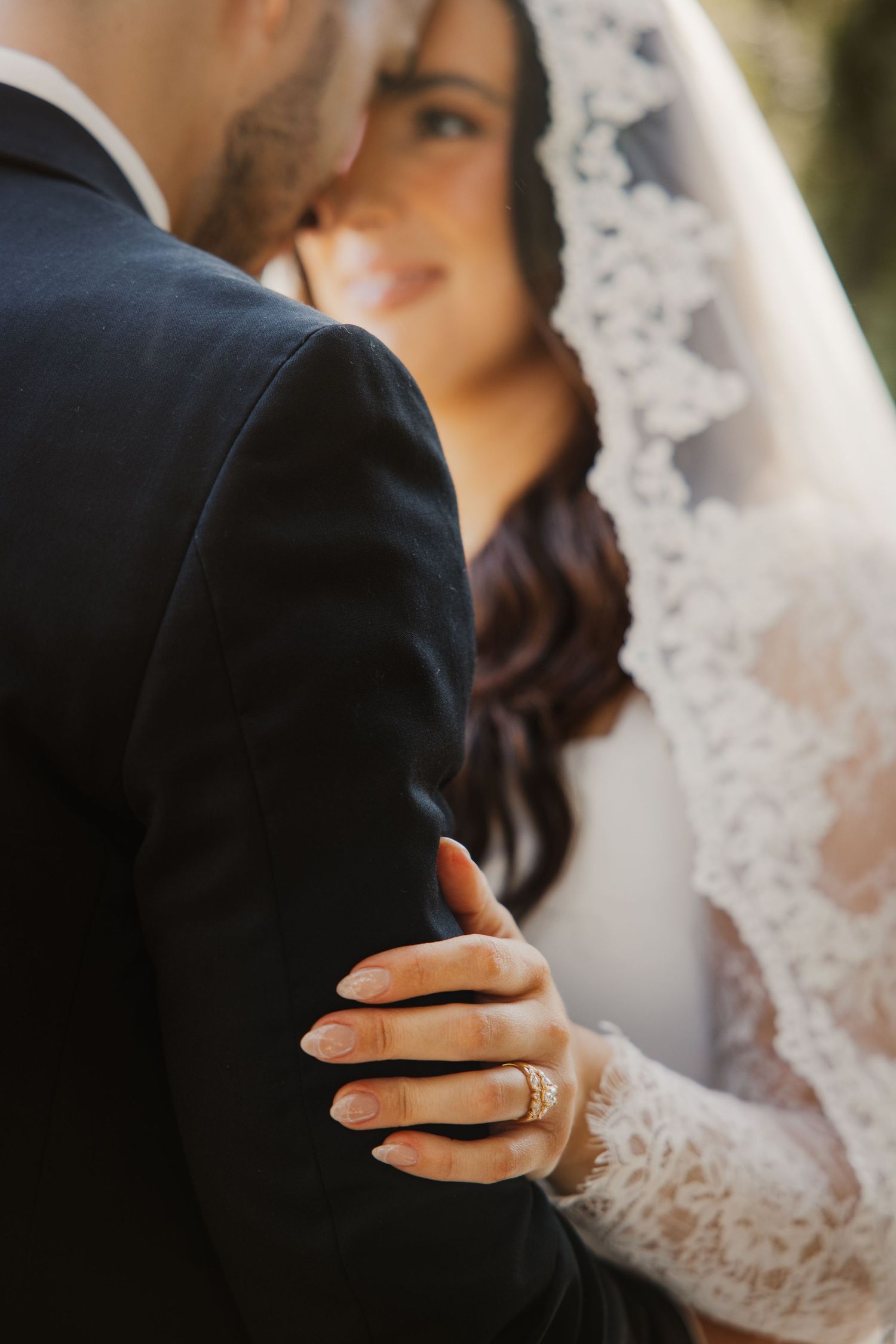 Close-up of bride in lace wedding dress with ring visible as she embraces groom outdoors.