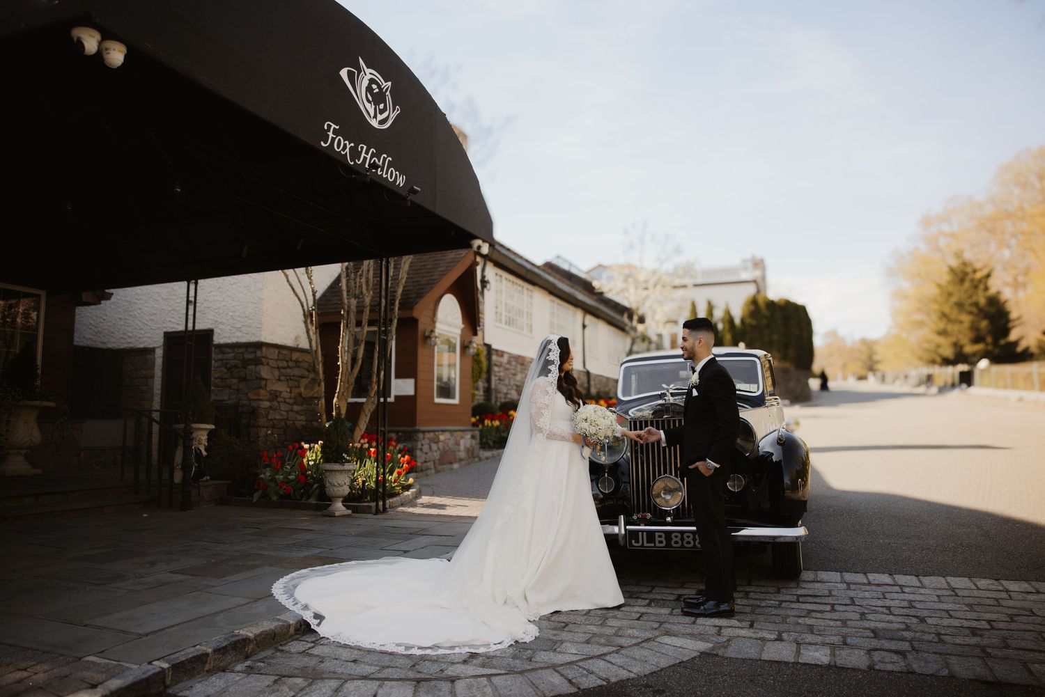 Bride and groom pose beside a vintage black car outside an elegant venue with a dark awning on a sunny day.