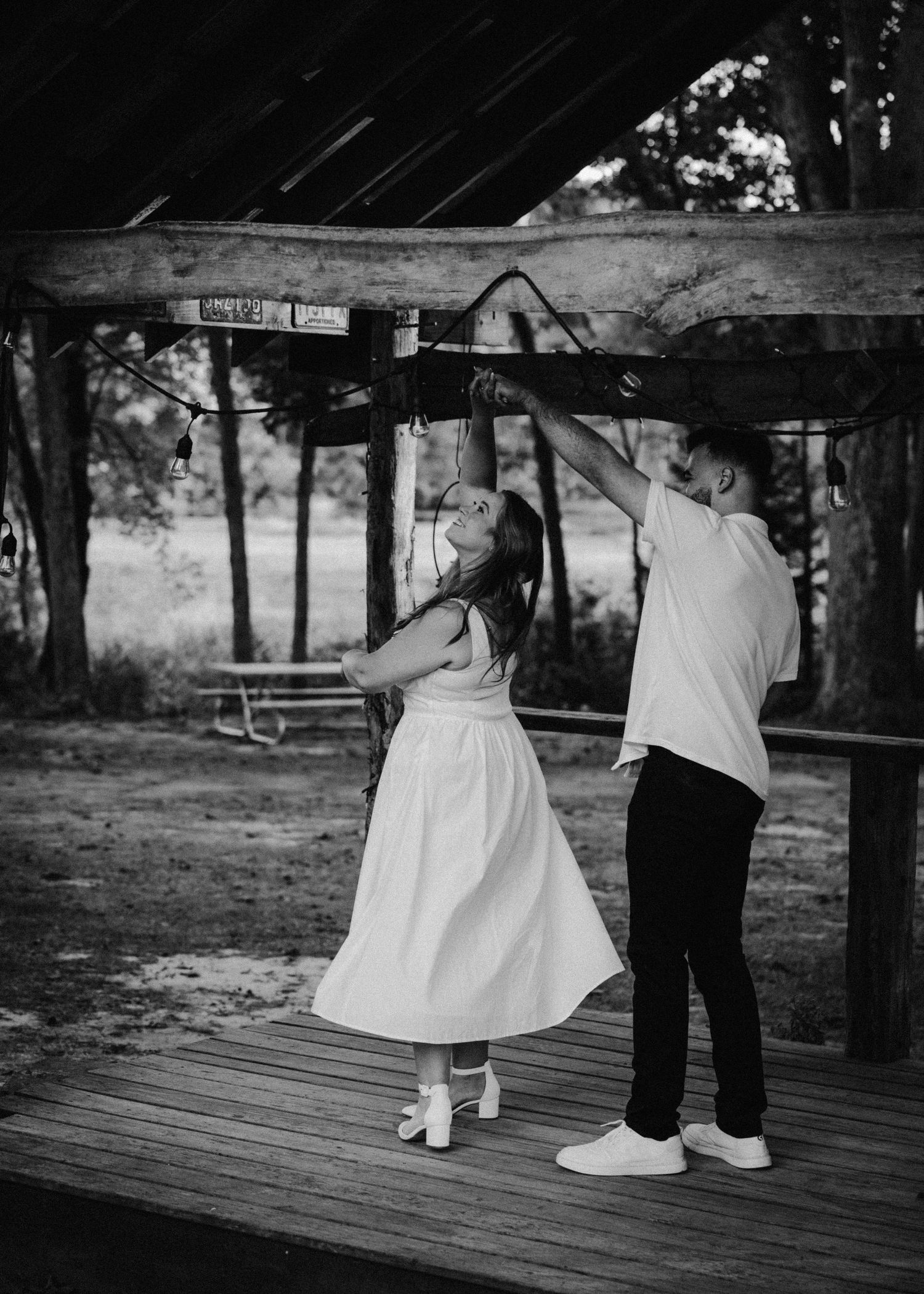 A couple dances playfully under a rustic wooden pergola in a black and white photo.