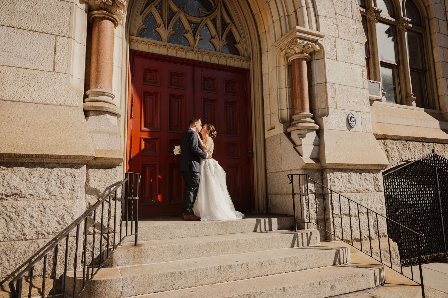 Bride and groom share a romantic kiss on the steps of a grand stone church with red doors.