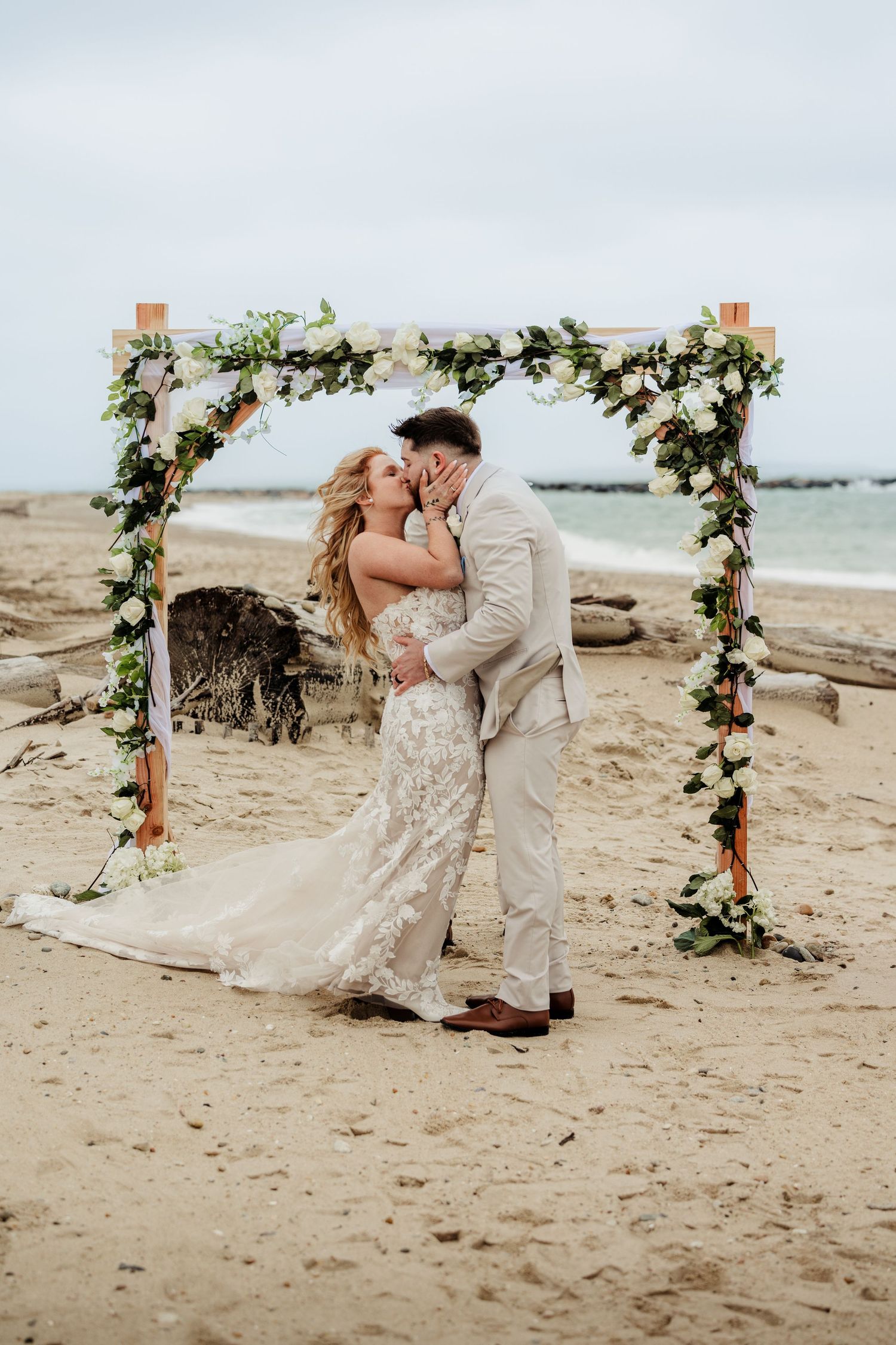 Bride and groom share a kiss under a floral arch during a romantic beach wedding ceremony.