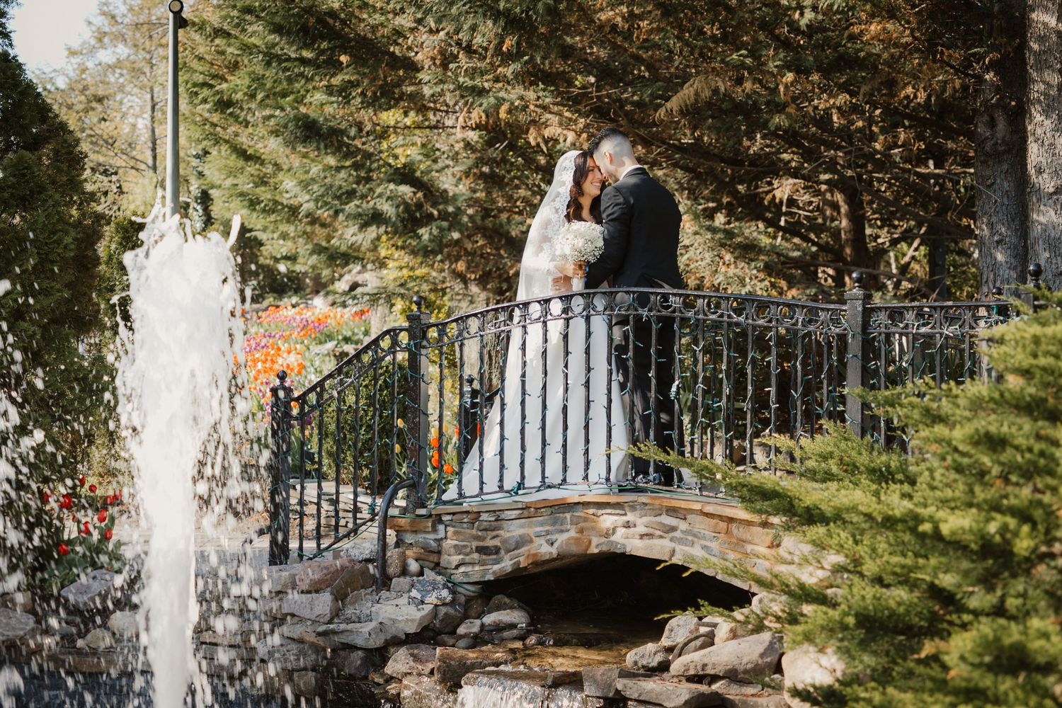 Bride and groom share a romantic kiss on a stone bridge with a fountain and autumn trees in a garden wedding setting.