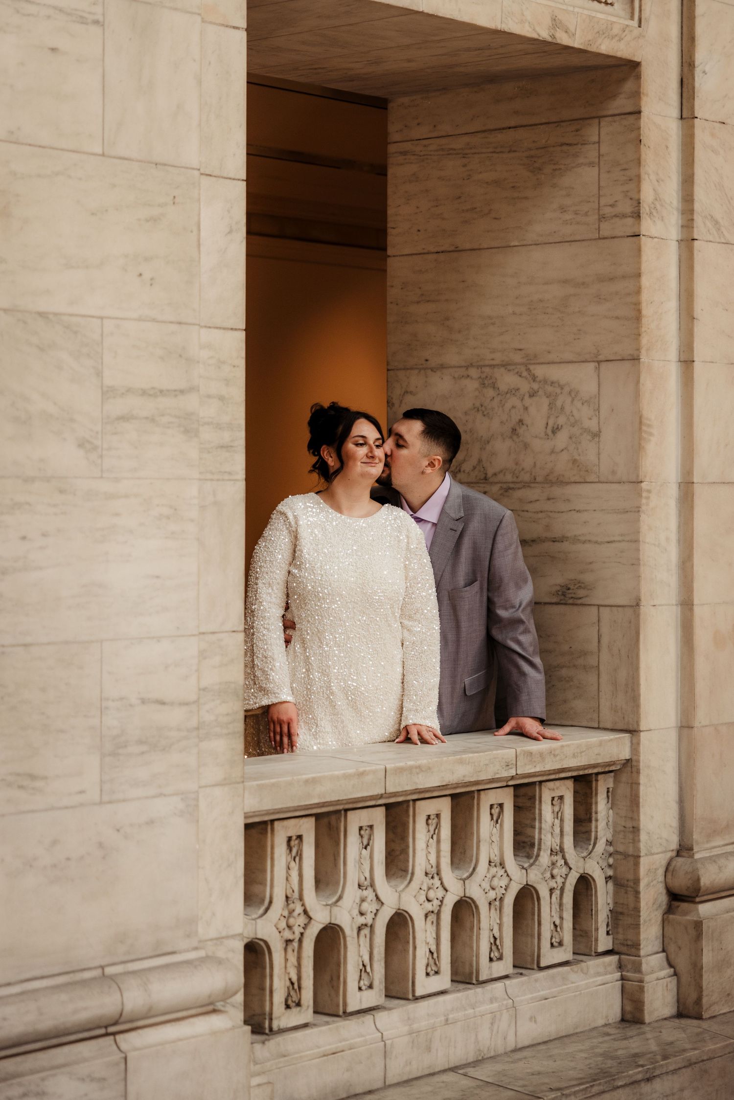 Groom kisses bride in white sequin dress while standing in marble architectural alcove.