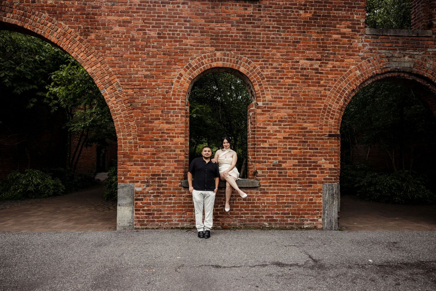 Couple poses at brick archway, woman sitting in arch while man stands below during outdoor engagement shoot.
