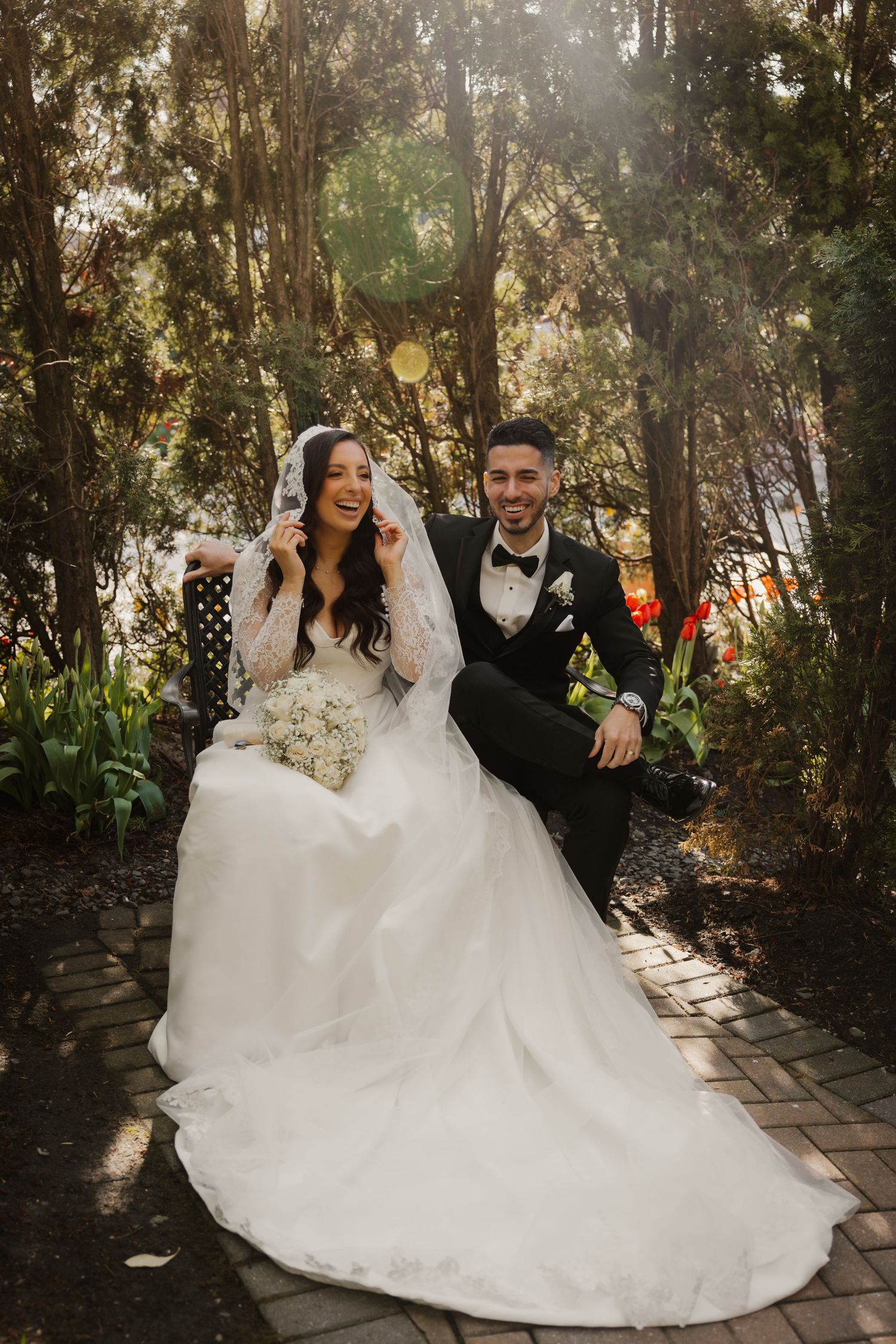 Bride and groom sitting on garden bench, bride in white gown with veil, groom in black tuxedo, surrounded by trees.