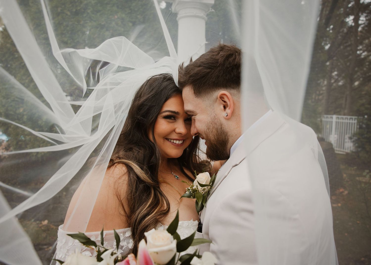 Bride and groom share intimate forehead touch moment under flowing veil with bridal bouquet.