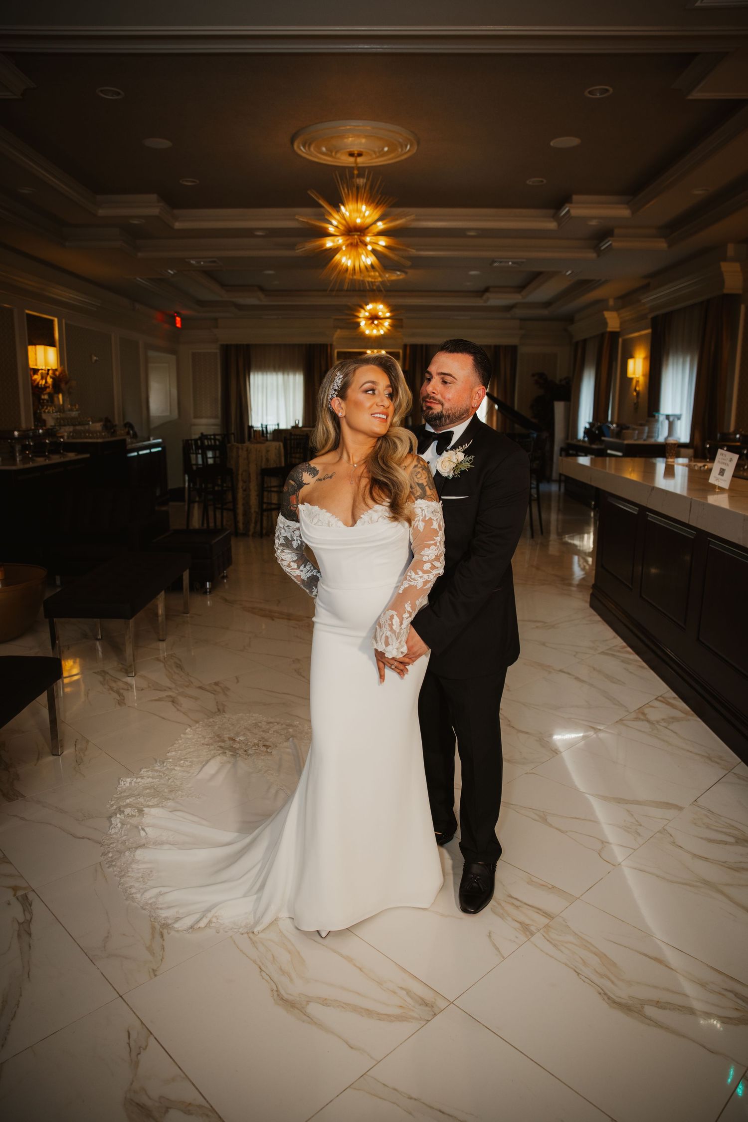 Bride in lace sleeve mermaid gown and groom embracing in luxurious marble ballroom.