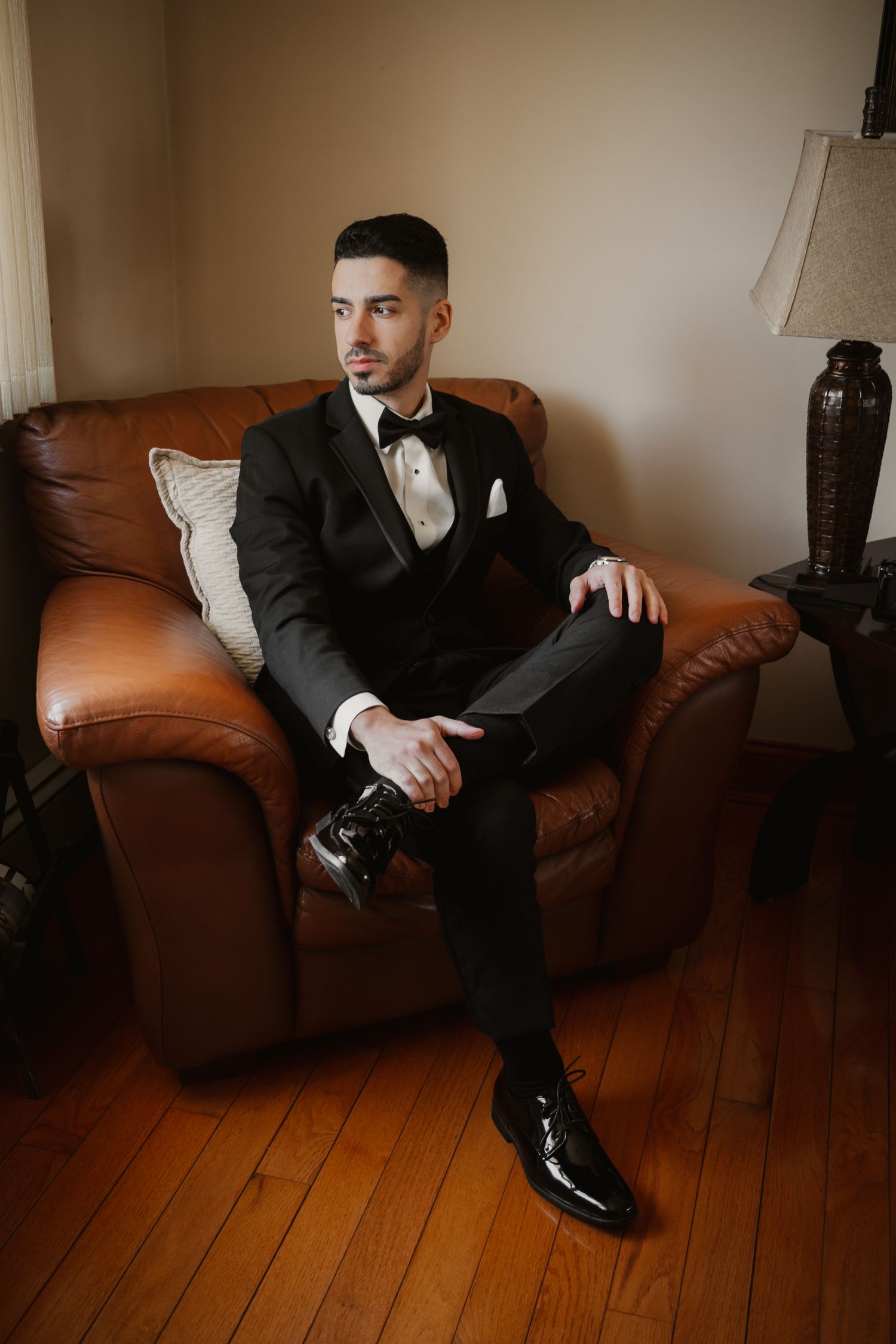 Elegant groom in black tuxedo with bow tie sits confidently in a leather armchair indoors.