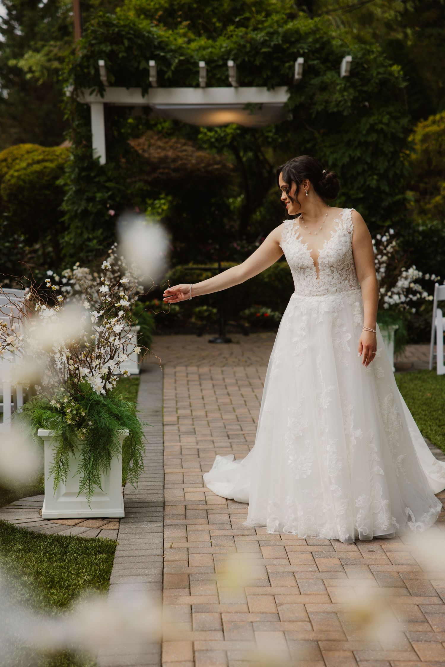 Bride in lace A-line gown reaches toward floral arrangement on garden wedding ceremony aisle.