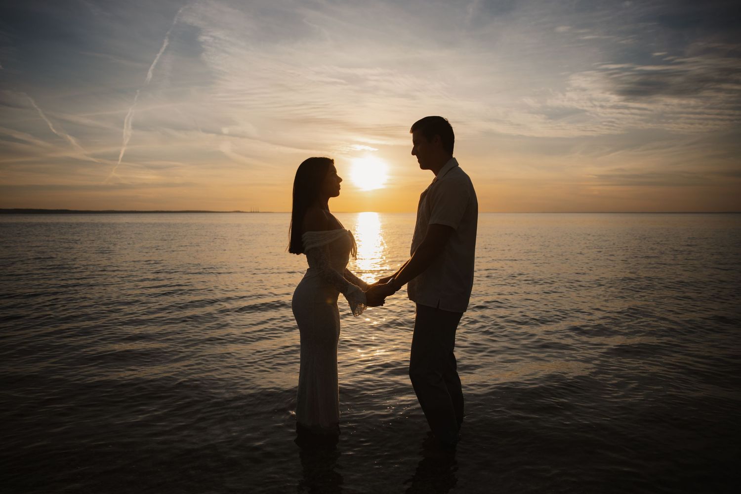 Romantic couple holding hands in silhouette against a golden sunset over calm ocean waters.