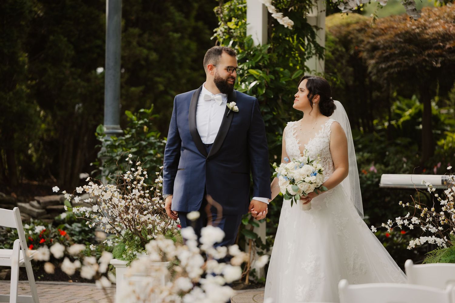 Bride and groom hold hands and smile at each other during outdoor wedding ceremony surrounded by lush greenery and white florals.