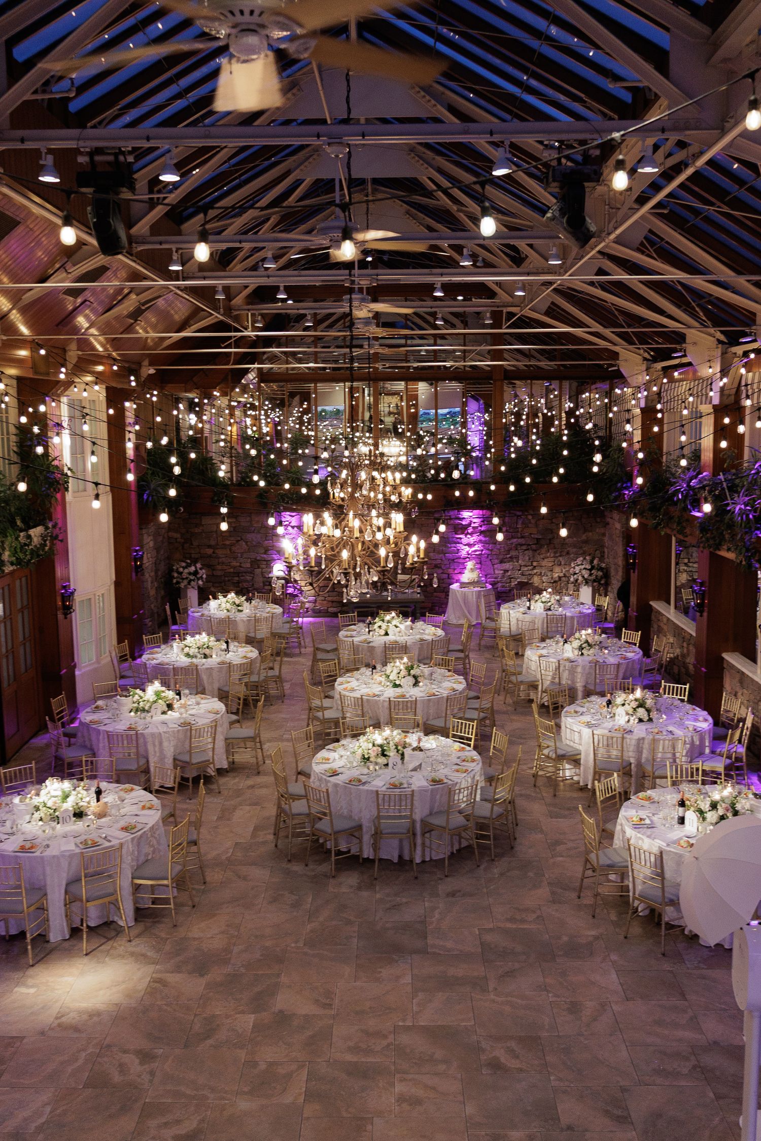 Elegant wedding reception hall with round tables, fairy lights, chandelier, and purple ambient lighting in a rustic barn venue.
