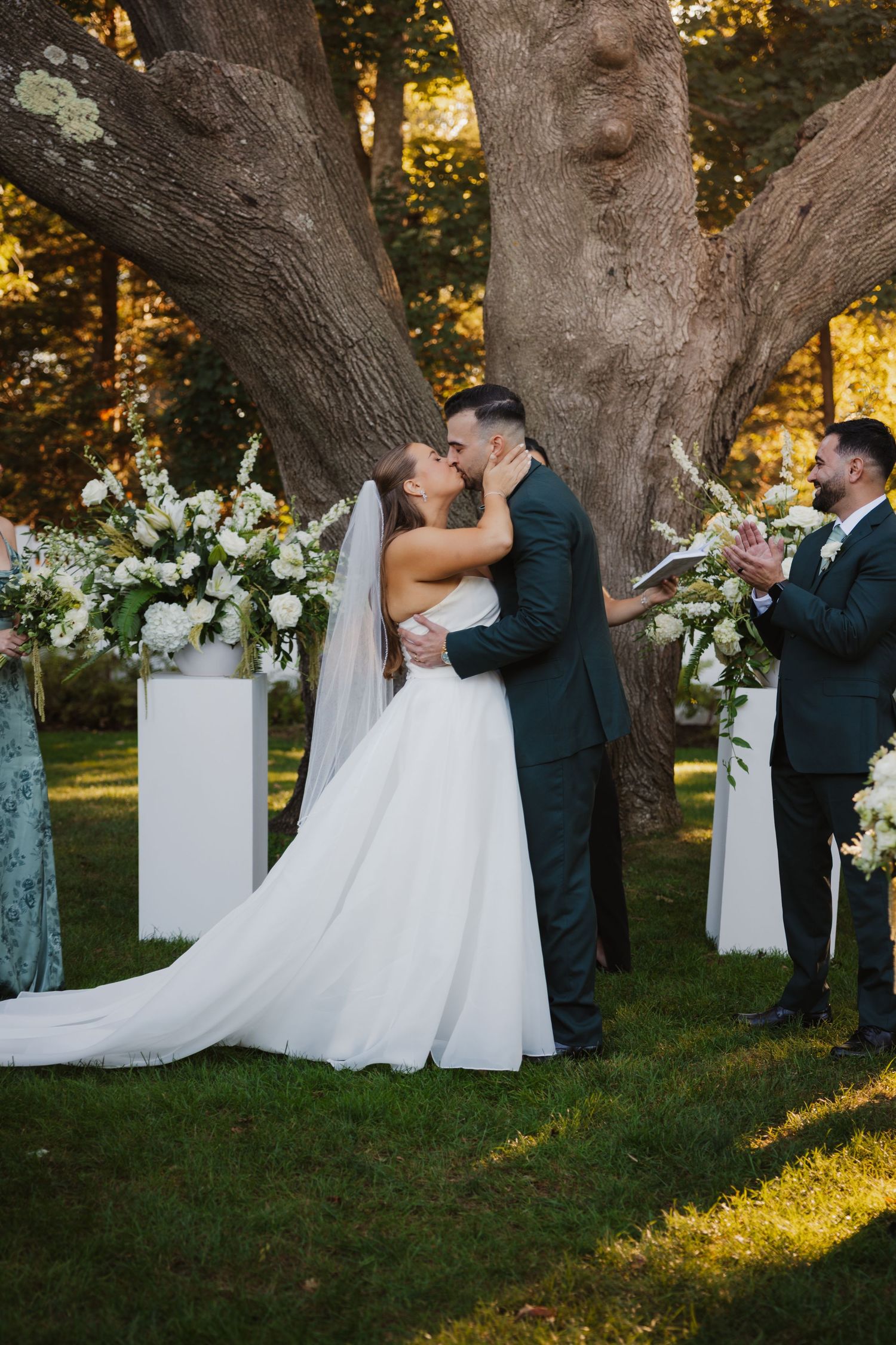 Bride and groom share first kiss at outdoor wedding ceremony under large tree with white floral arrangements.