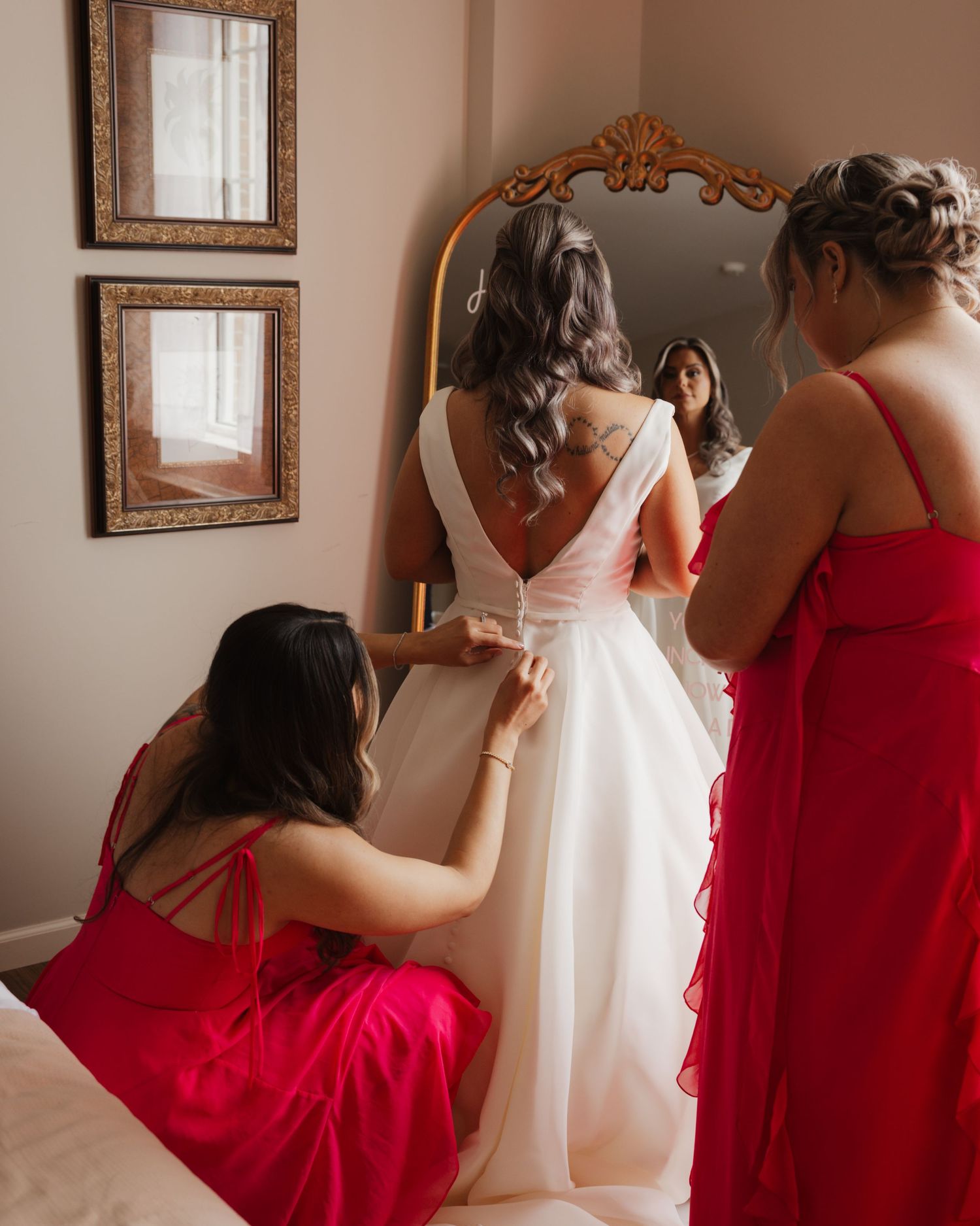 Two bridesmaids in red dresses help bride with her white gown in front of ornate gold mirror.