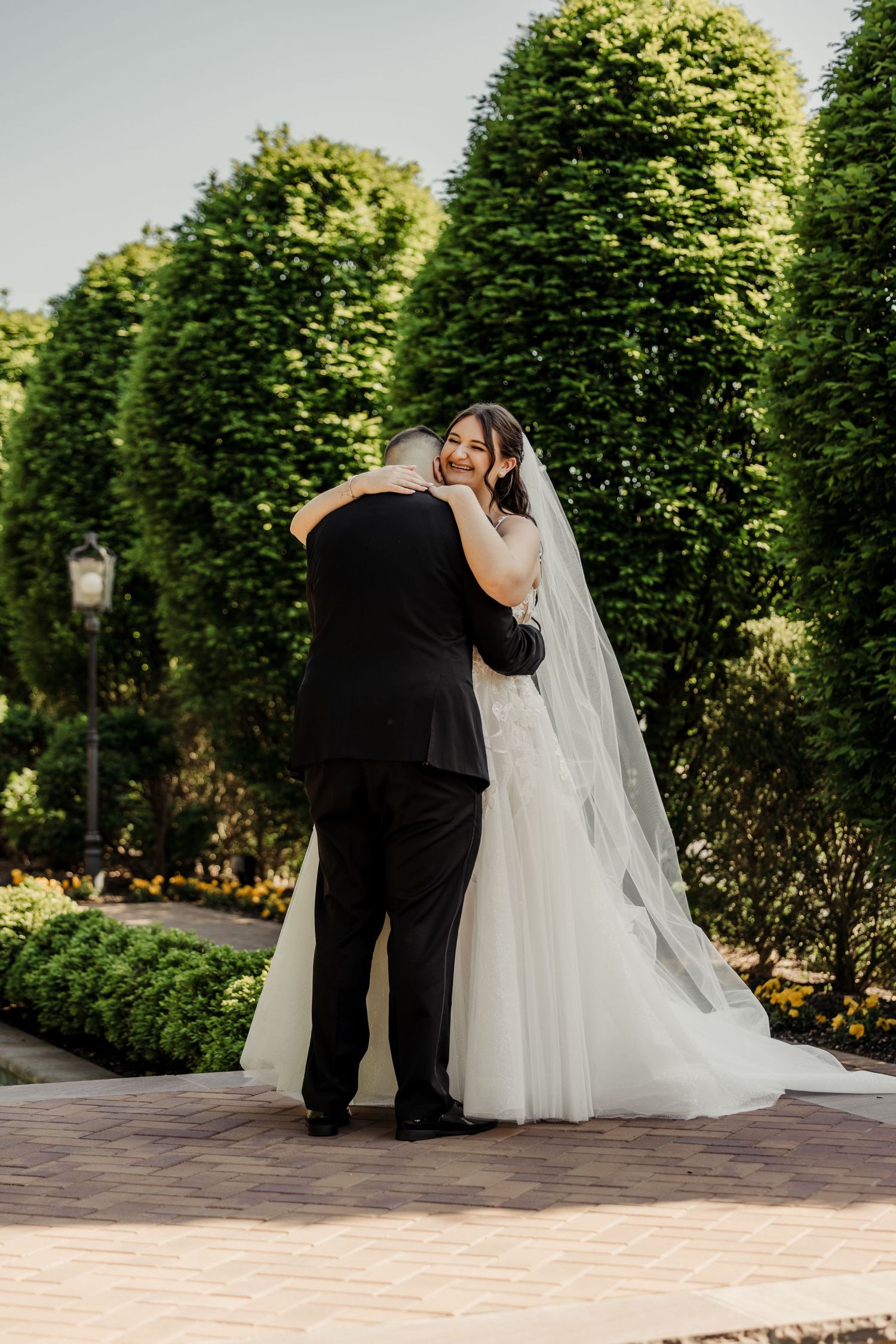Bride in a flowing white gown and veil embraces groom on a brick garden path surrounded by trees.