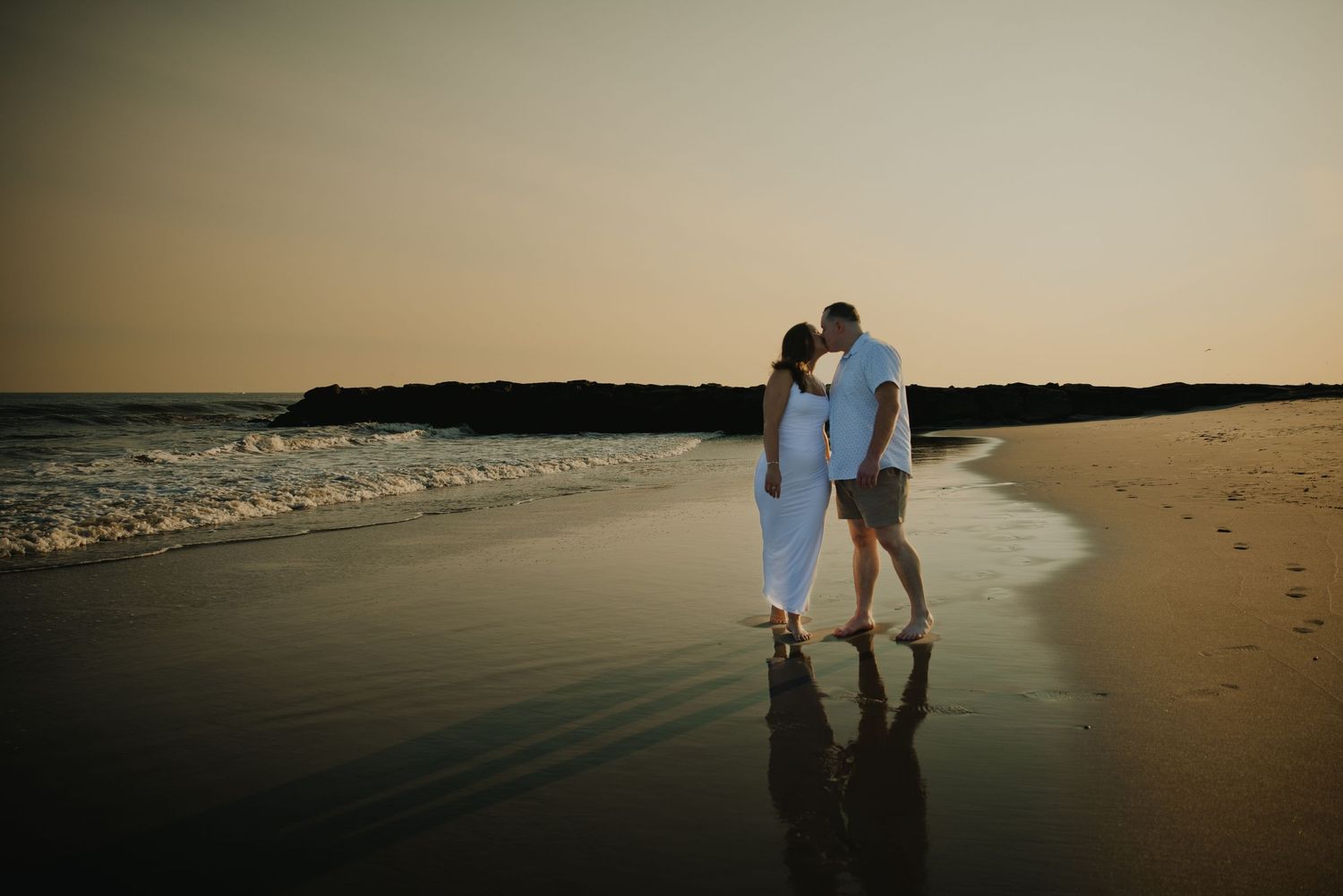 Newlyweds kissing on a beach at sunset with waves reflecting golden light.