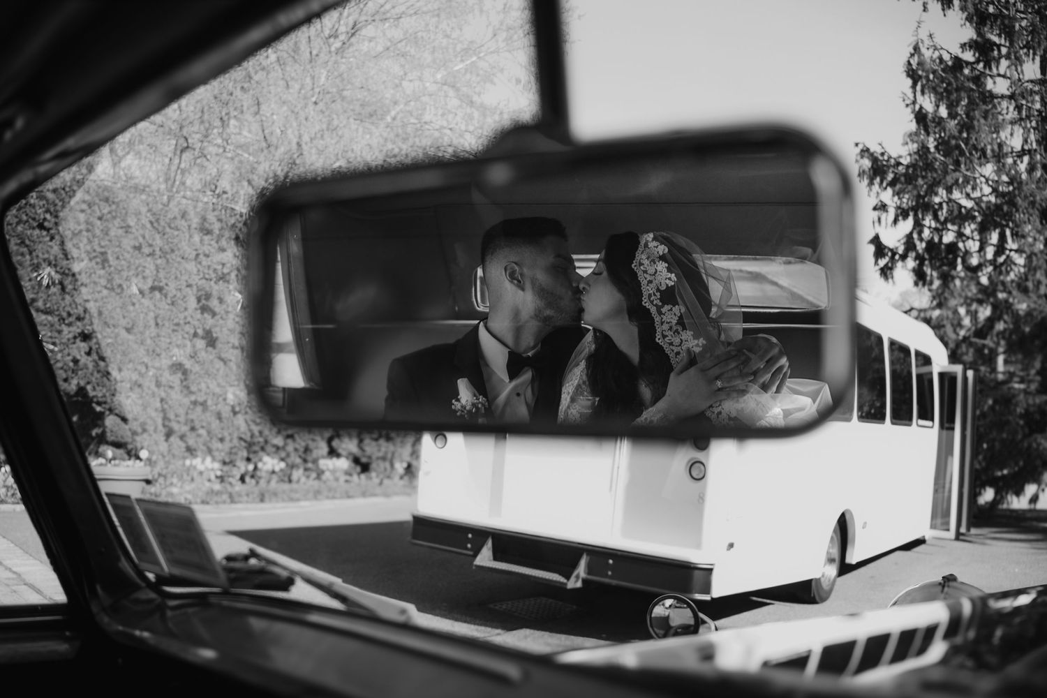 Black and white photo of a couple kissing reflected in a vintage car rearview mirror with a bus visible outside.