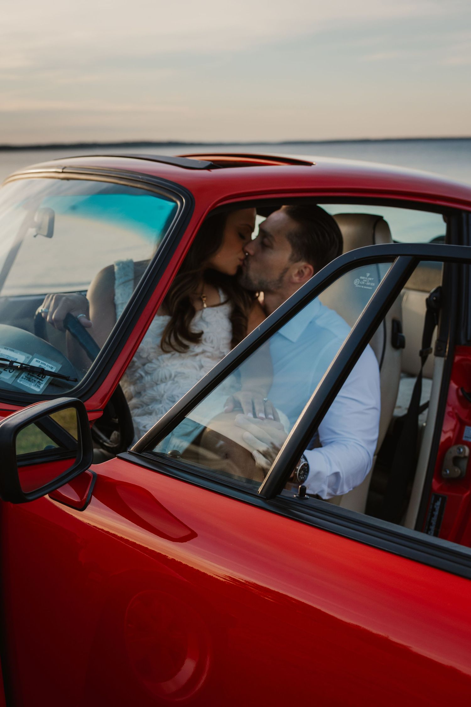 Couple sharing a romantic kiss inside a classic red vintage car near a scenic waterfront.