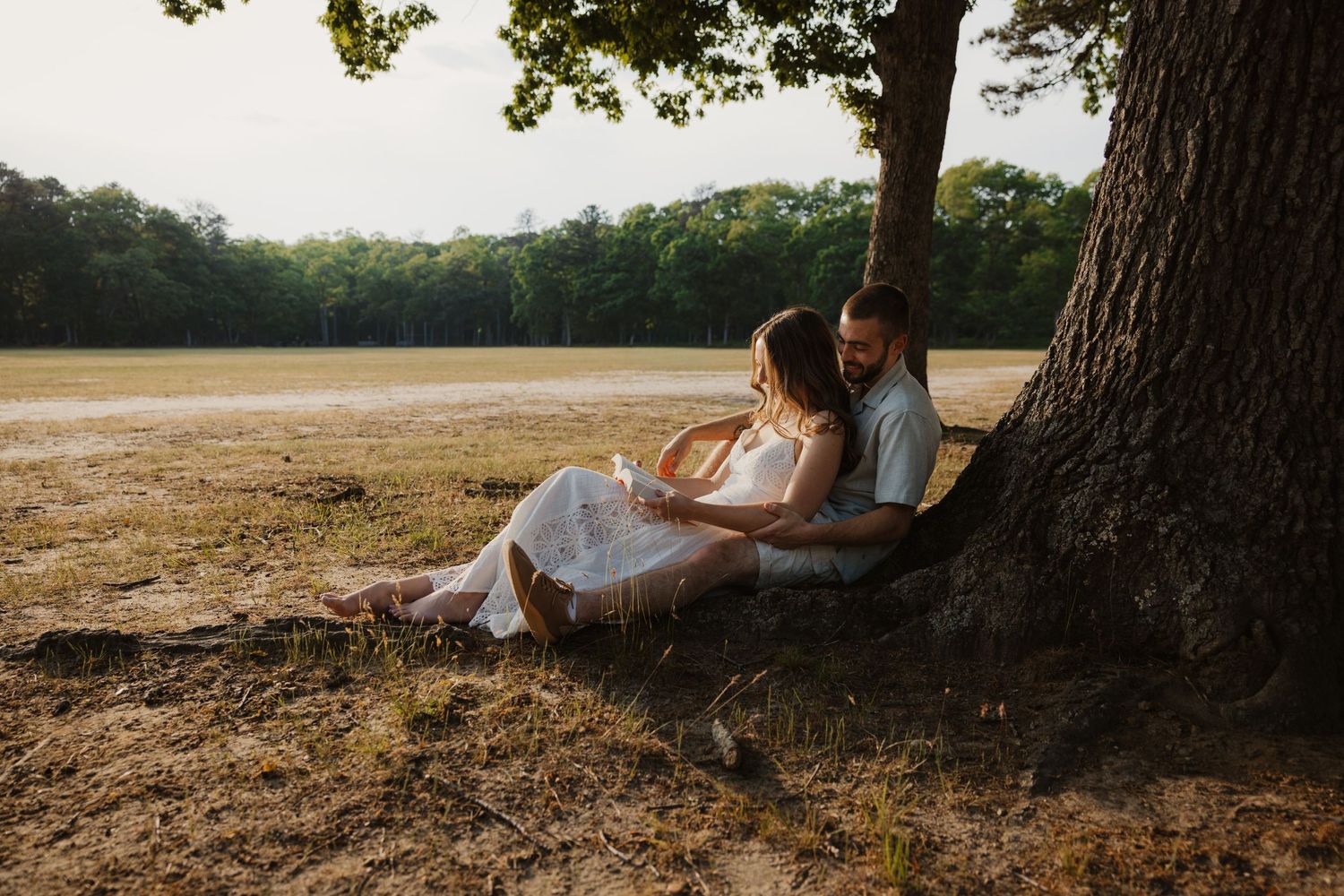 A couple sits leaning against a large tree trunk in a dry sunlit field, sharing a quiet moment.
