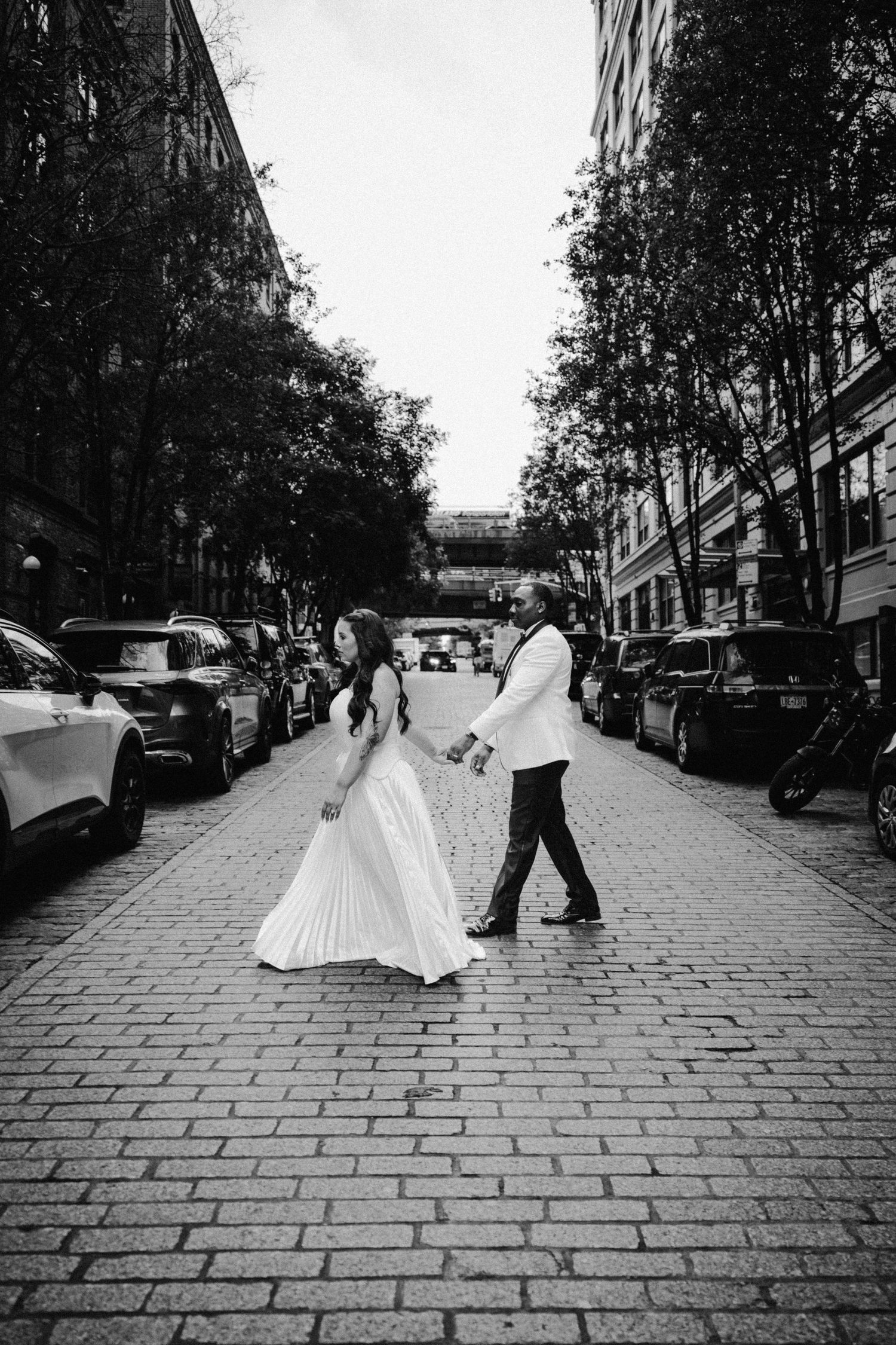 Black and white photo of bride and groom walking hand in hand across a cobblestone city street.