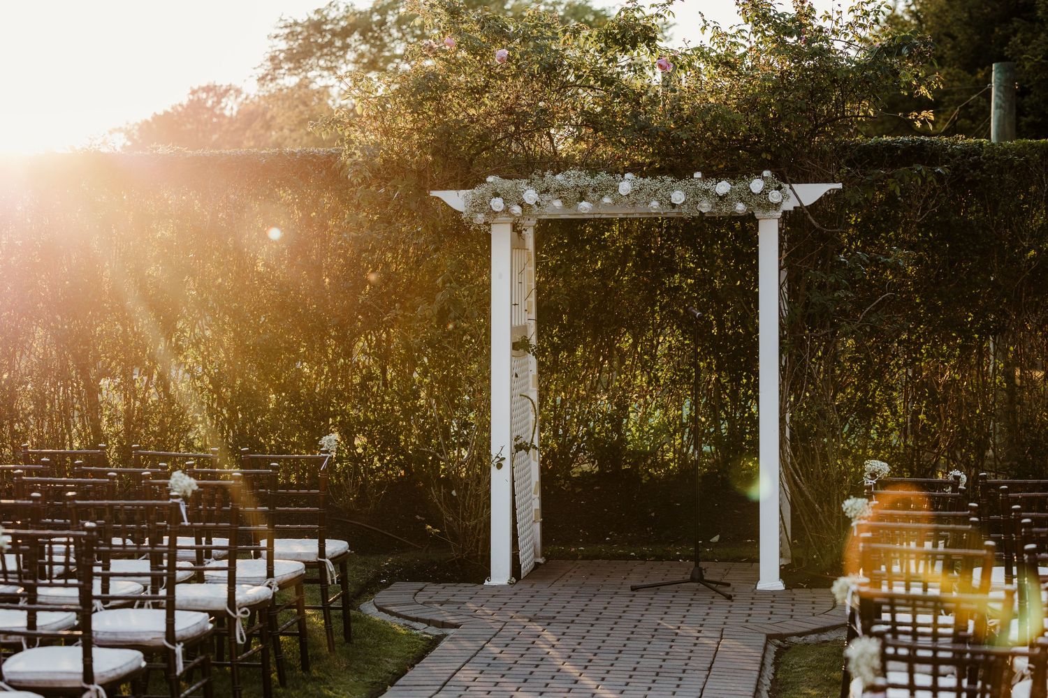 Sunlit white wedding arch adorned with flowers in a garden ceremony space with rows of chairs.