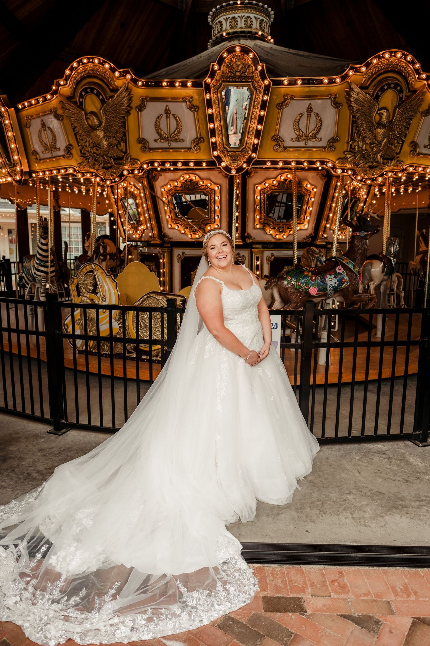Bride in full ballgown with lace veil poses elegantly in front of a lit vintage carousel.
