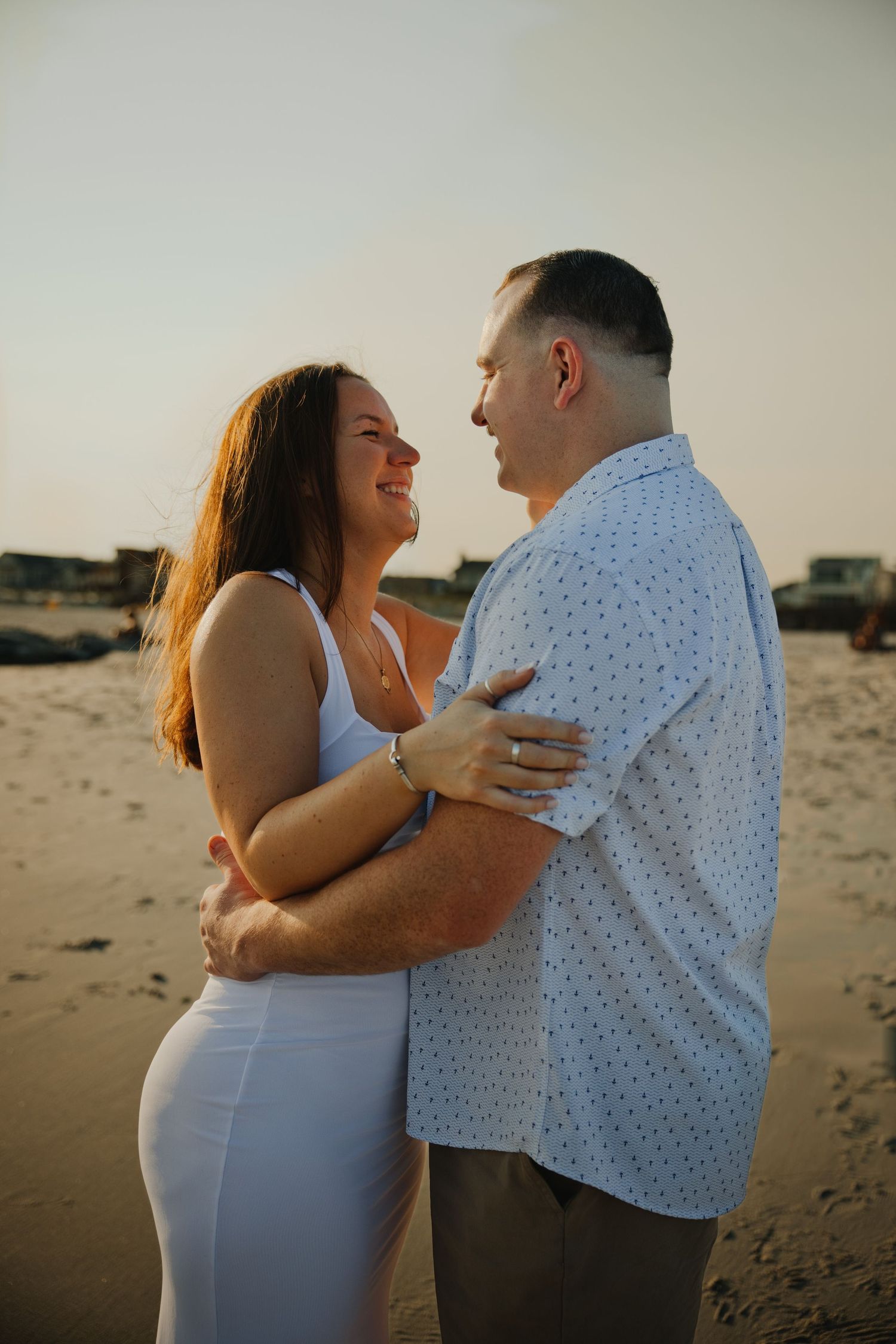 Smiling couple embracing on sandy beach at sunset, woman in white dress and man in blue shirt.