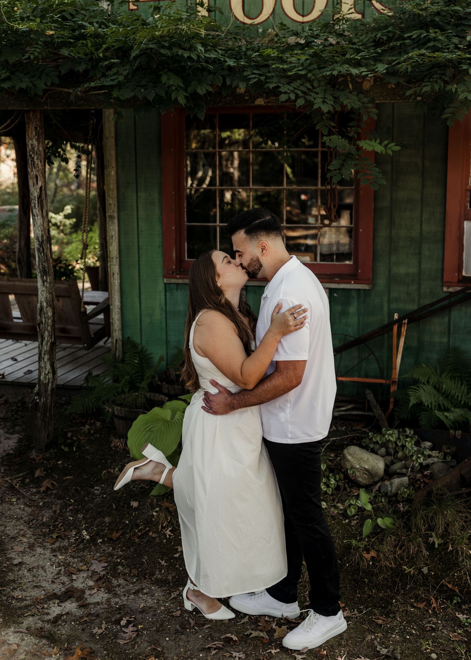A couple embraces and kisses outside a charming green cottage surrounded by lush greenery.
