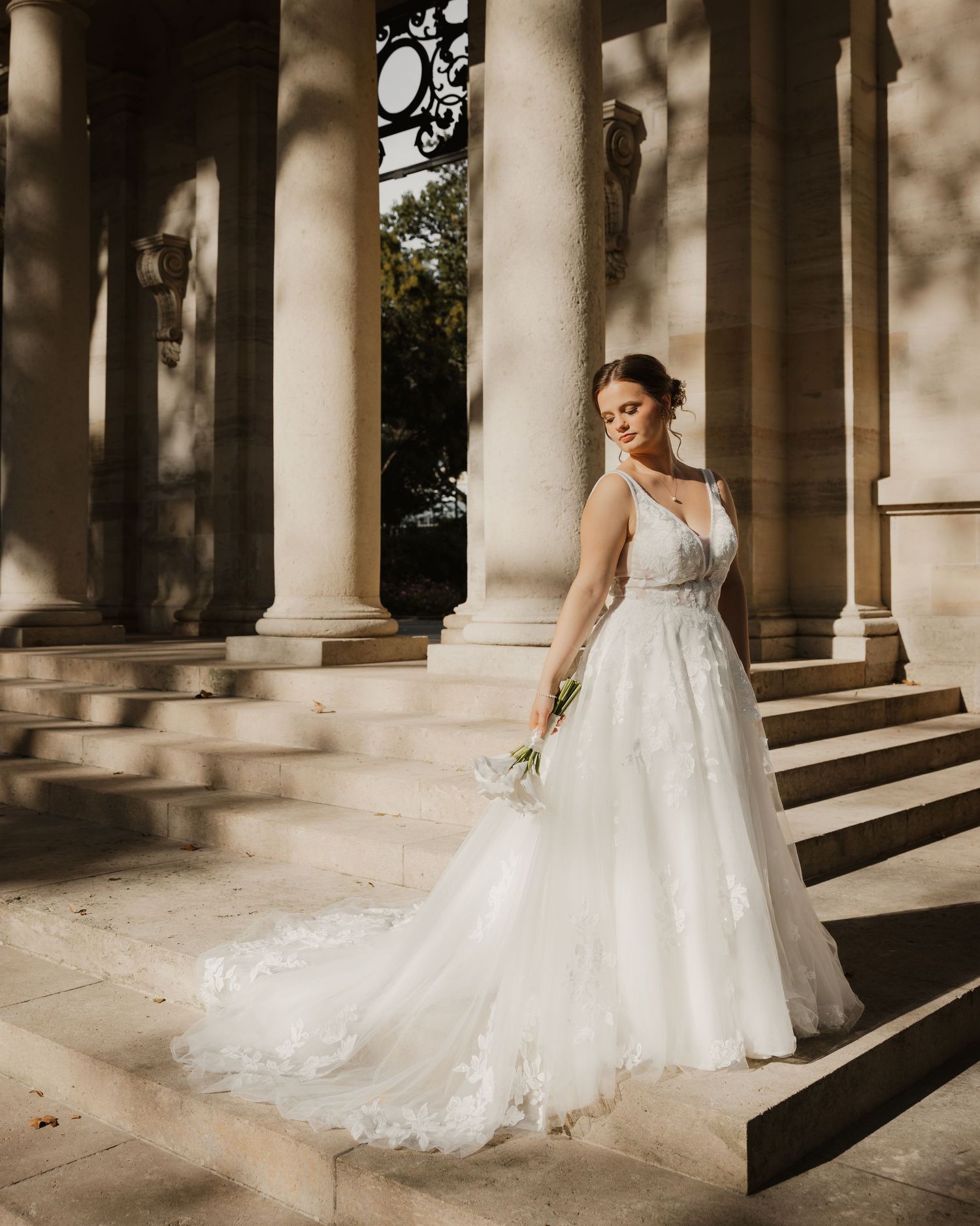 Bride in elegant lace A-line wedding gown posing on stone steps between classical columns.
